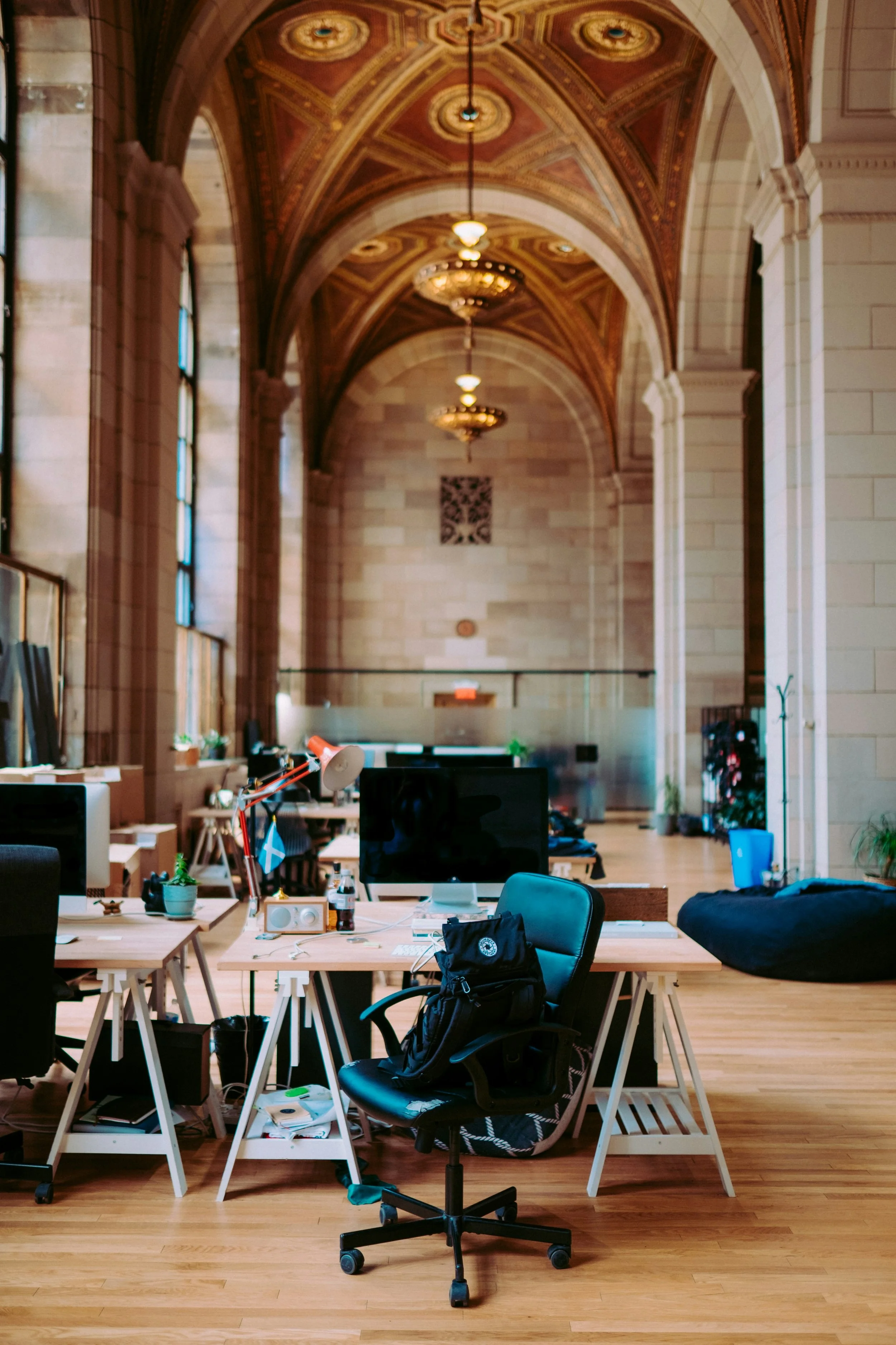 Open office workspace with multiple desks, chairs, computers, and office supplies inside a building with high, ornate ceilings and large windows.