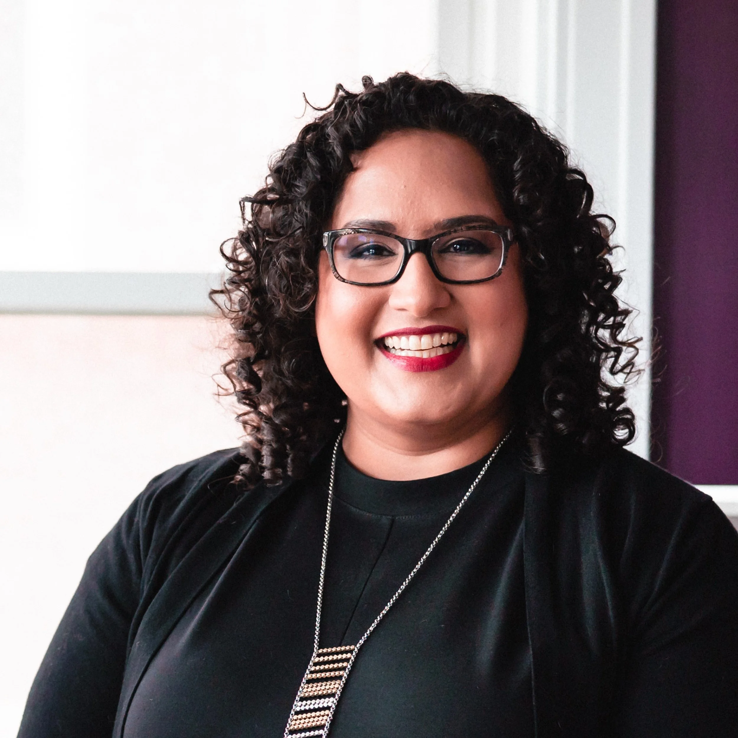 Close-up of woman with curly hair, wearing black-rimmed glasses, red lipstick, leather jacket, and a silver chain necklace, smiling in front of a light background.