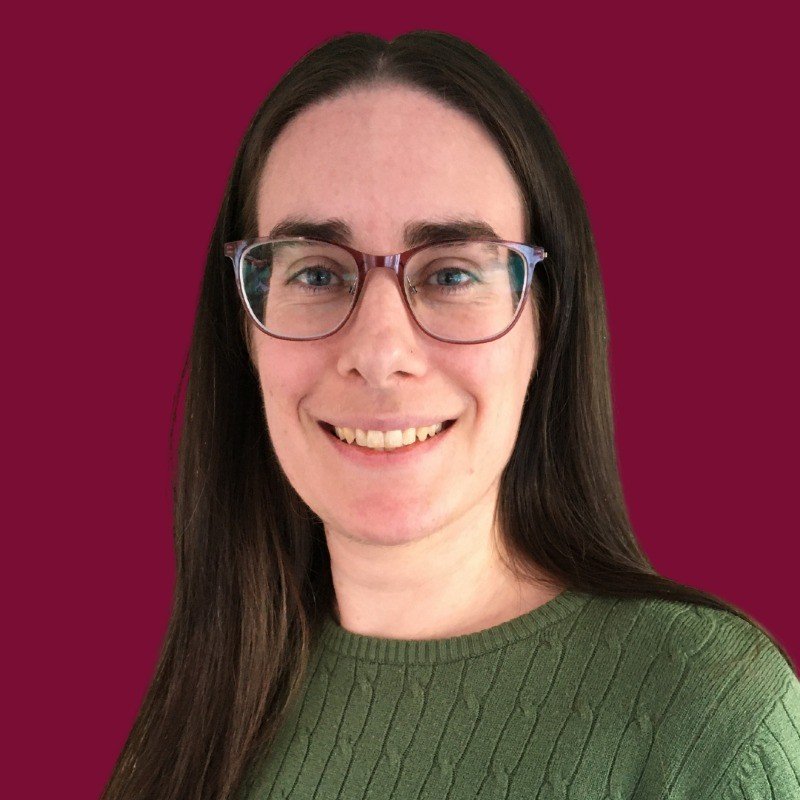 A woman with long dark hair, wearing glasses and a green sweater, smiling against a burgundy background.