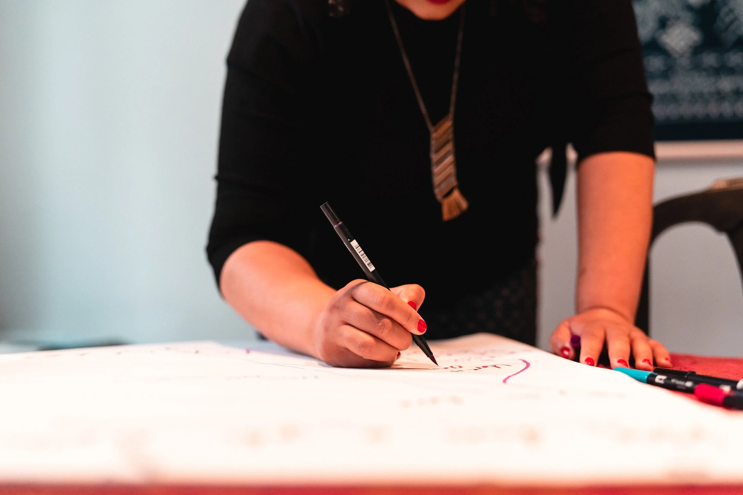 A person with red painted nails writing on a large sheet of paper with a black marker, using their right hand. The person is wearing a black top and a patterned necktie, and is leaning over the table. The background is plain with light-colored walls, and a few colorful markers are seen on the table.