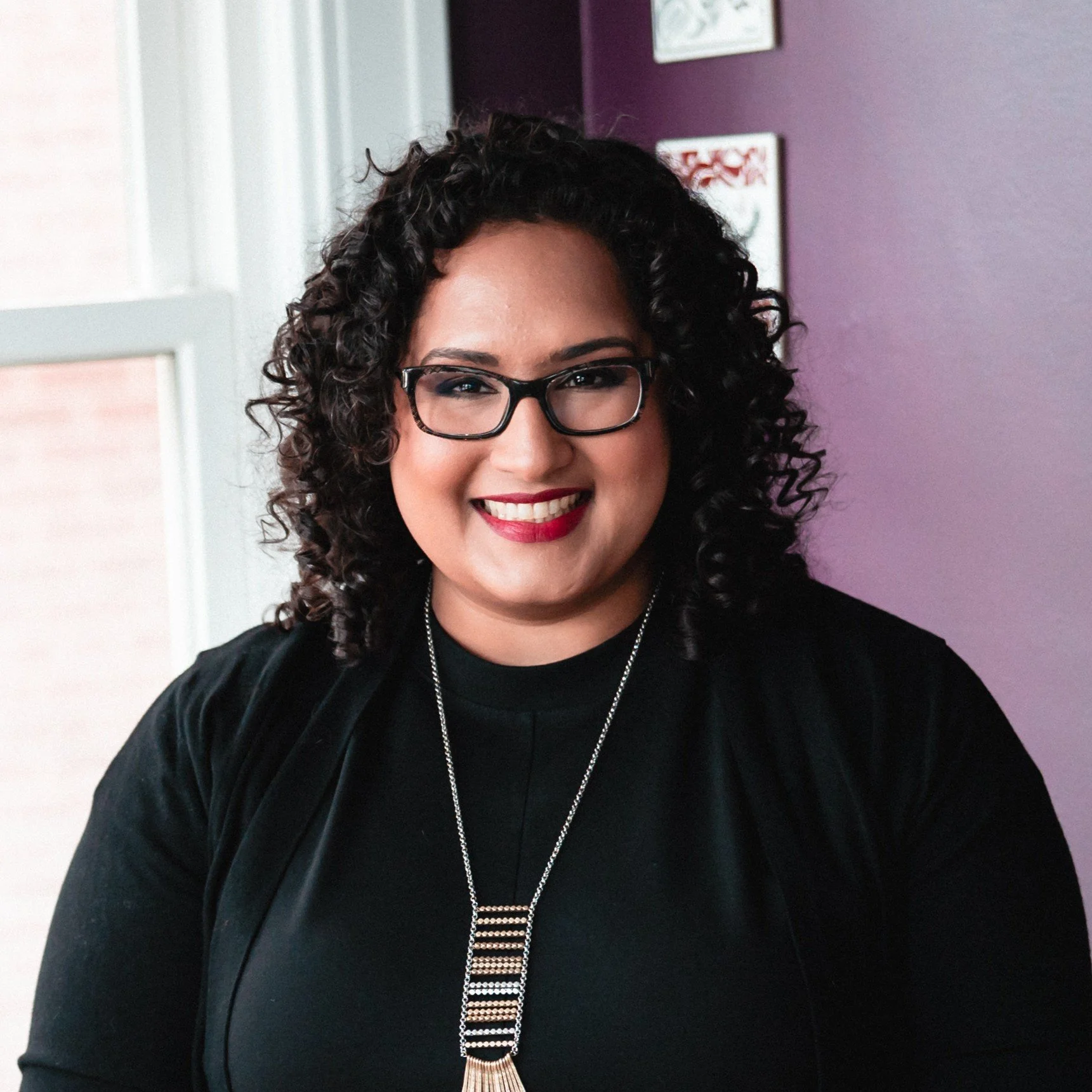 A woman with curly dark hair, glasses, red lipstick, and a black top, standing indoors against a purple wall with framed artwork.