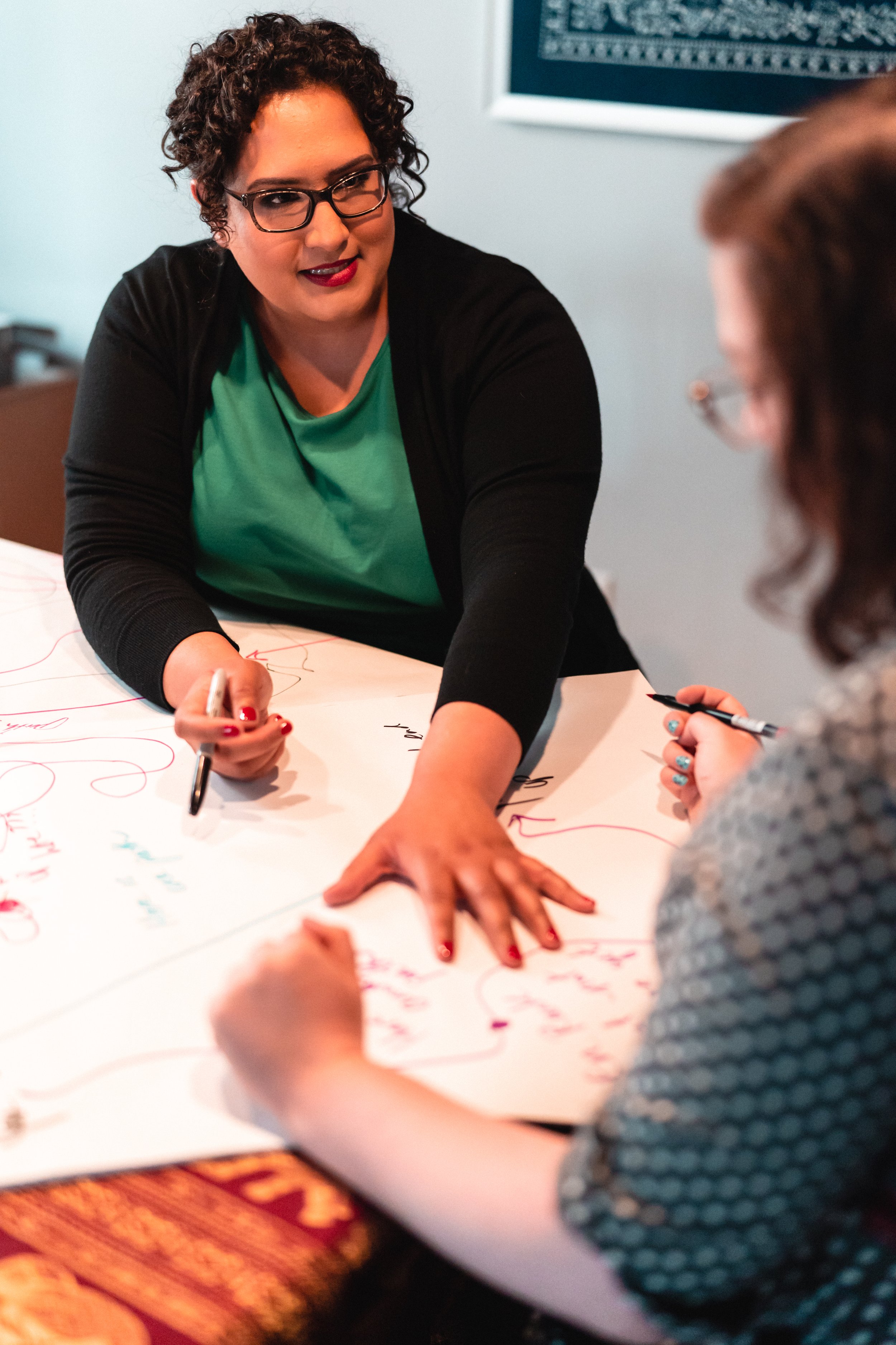 Two women are engaged in a discussion or brainstorming session, writing and drawing on a large sheet of paper spread across a table.