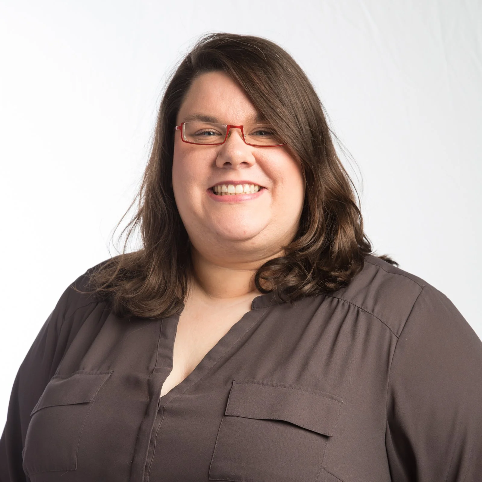 A woman with long brown hair wearing red glasses and a brown blouse, smiling against a plain light background.