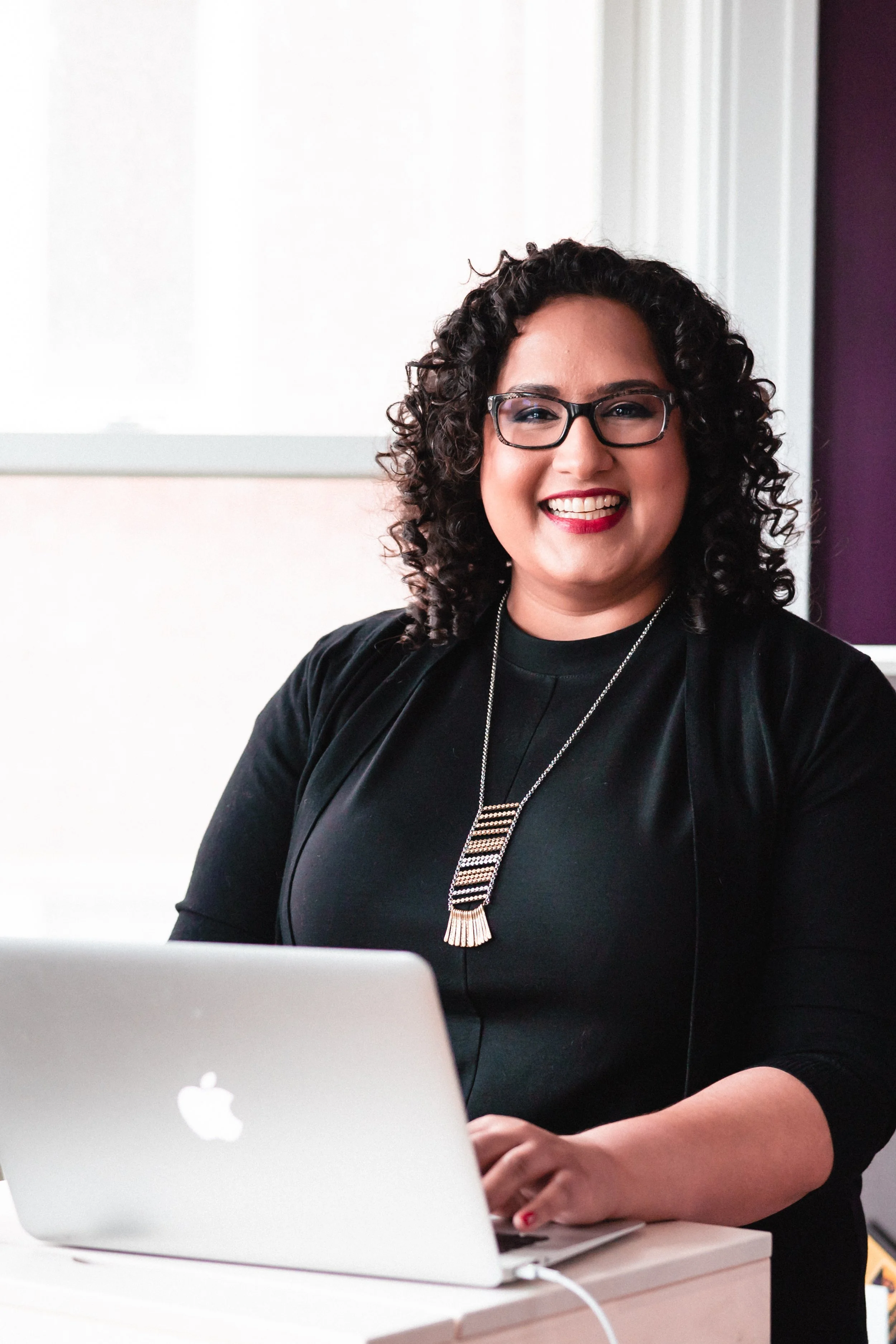 A woman with curly dark hair, glasses, wearing a black top and a necklace, smiling while working on a laptop at a desk.