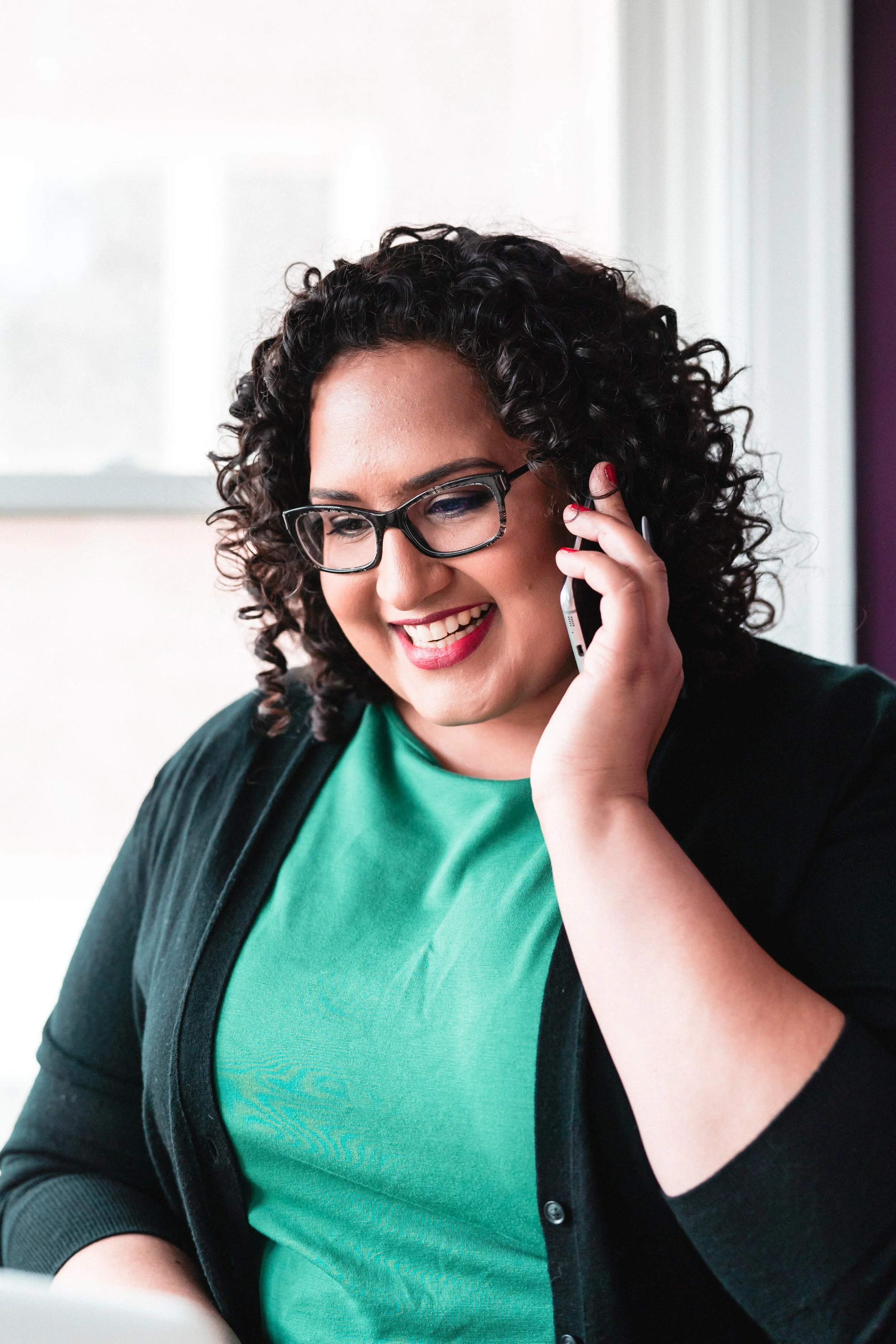 A woman with curly dark hair wearing glasses, a green shirt, and a black cardigan, smiling and talking on a mobile phone.
