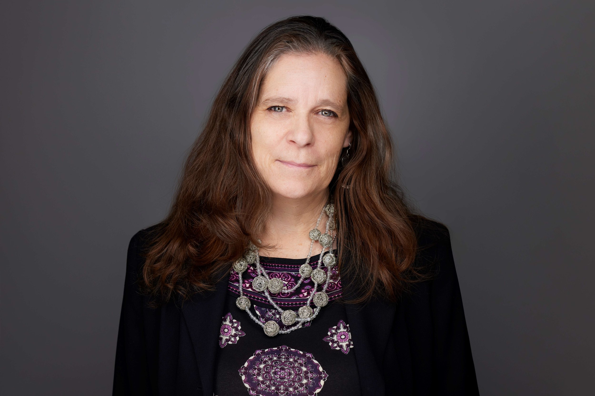 Portrait of a woman with long brown hair, wearing a black blazer and a purple patterned top, accessorized with layered silver necklaces and earrings, standing against a gray background.