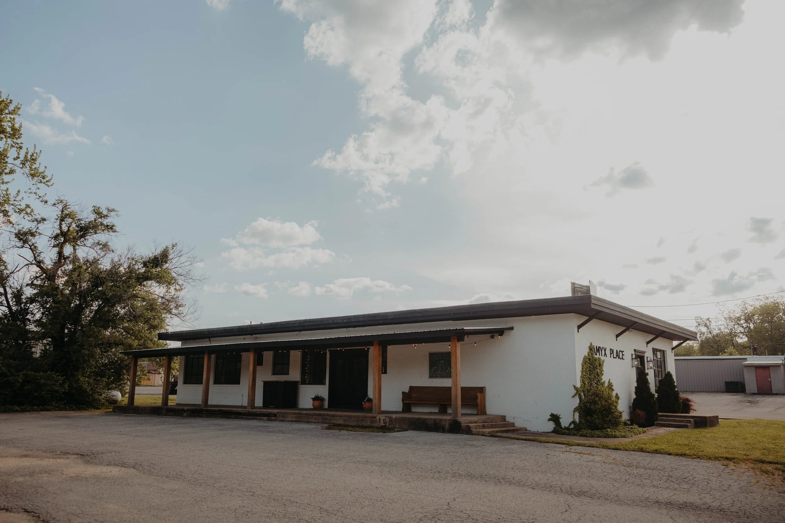 A white building with a dark roof and large windows, labeled 'AMYX PLACE.' The building has a small porch with wooden supports, a bench, and some potted plants outside. Trees and a gravel parking area are visible in front of the building, under a par