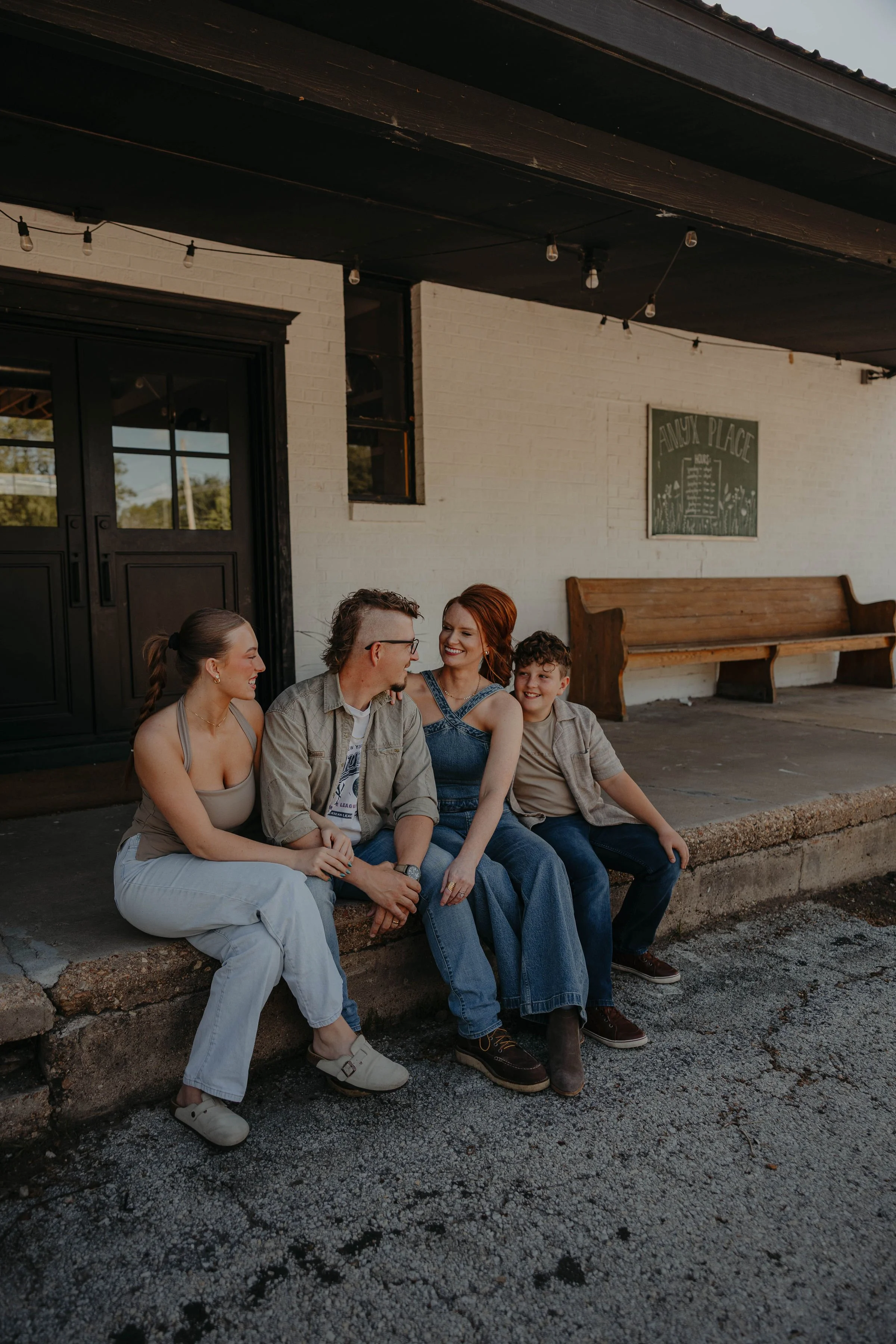 A group of five friends sitting on a sidewalk outside a building, engaged in conversation and smiling. The background shows a white brick wall with a sign that reads 'ANUKY PLACE' and a wooden bench.