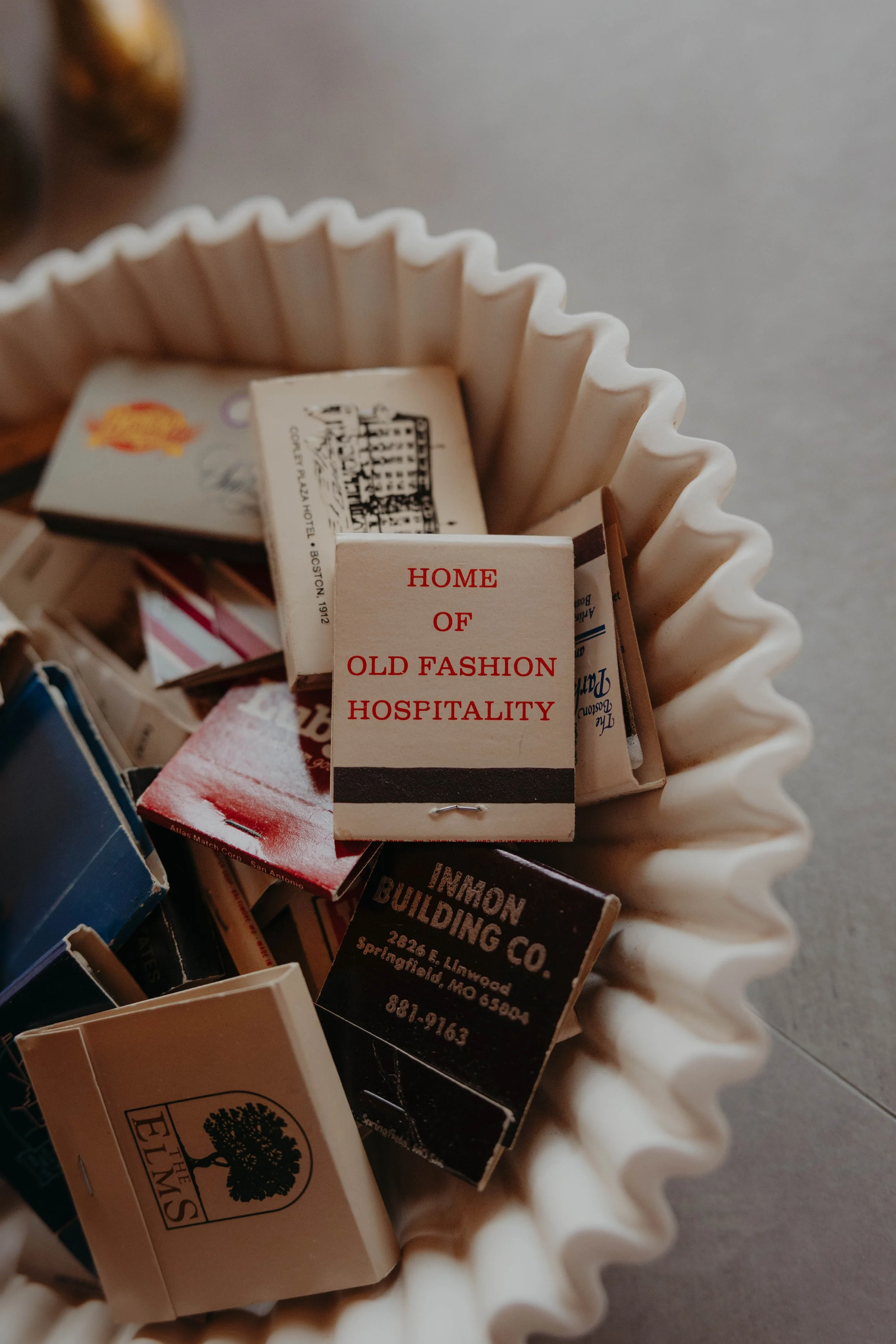A collection of matchbooks inside a white corrugated paper container, with one matchbook labeled "Home of Old Fashion Hospitality."