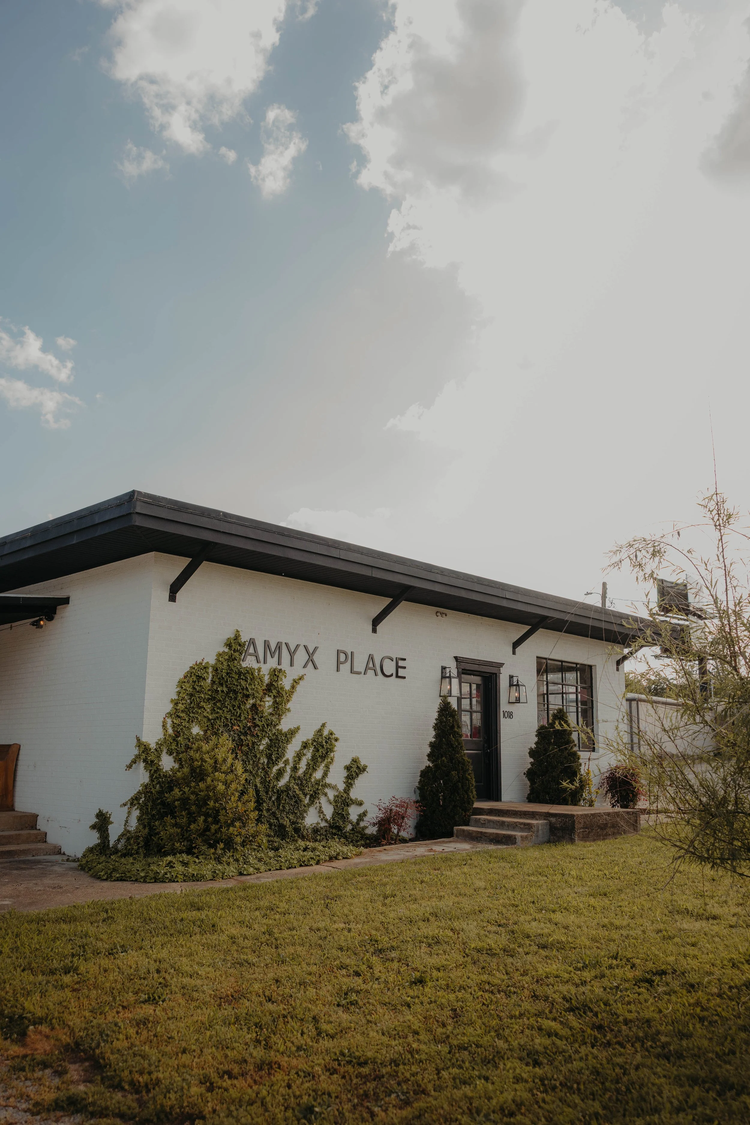 Exterior of a white building with black trim labeled 'AMYX PLACE,' surrounded by bushes and trees, under a partly cloudy sky.