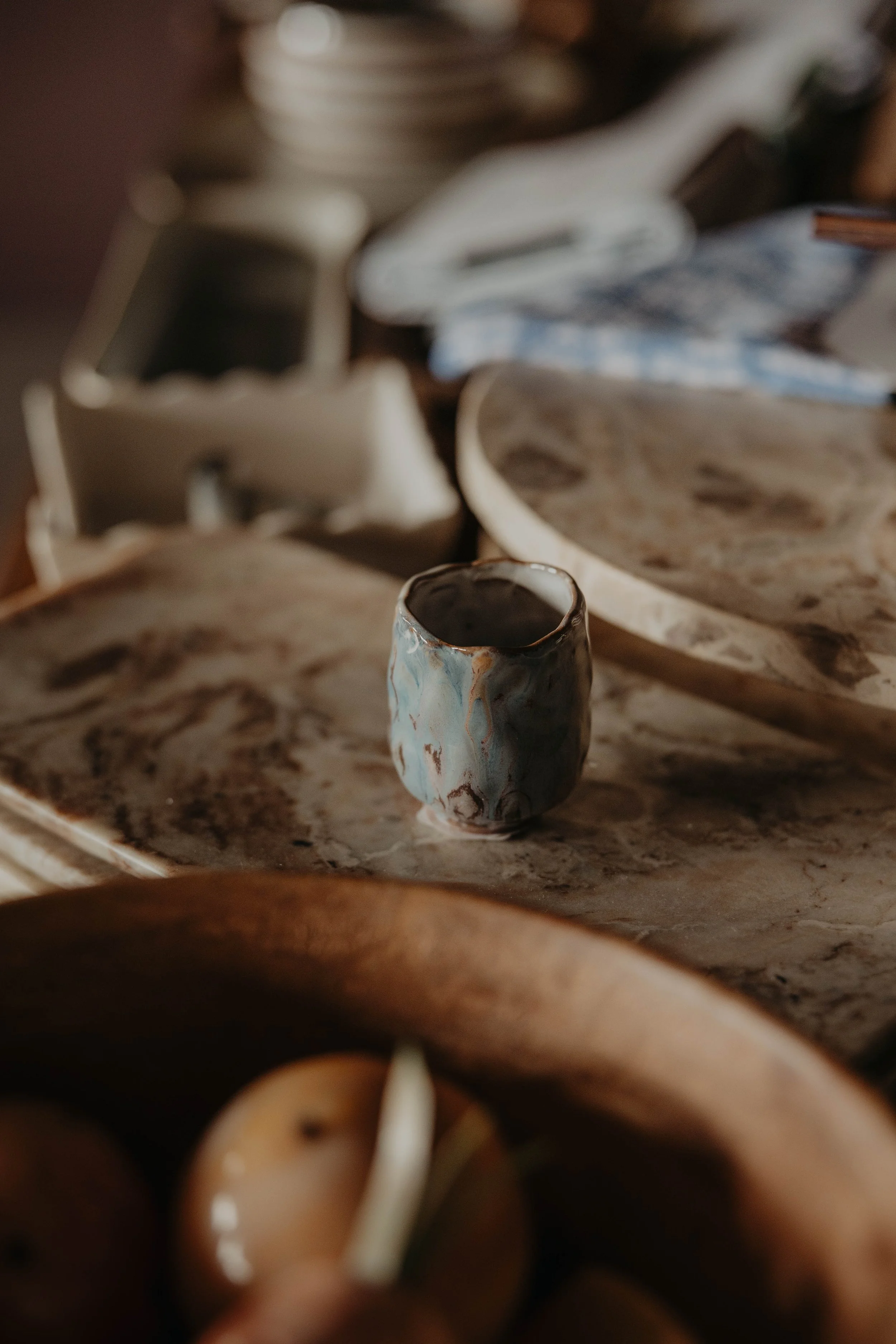 Close-up of a rustic ceramic cup on a marble surface, with blurred pottery and a blue cloth in the background.