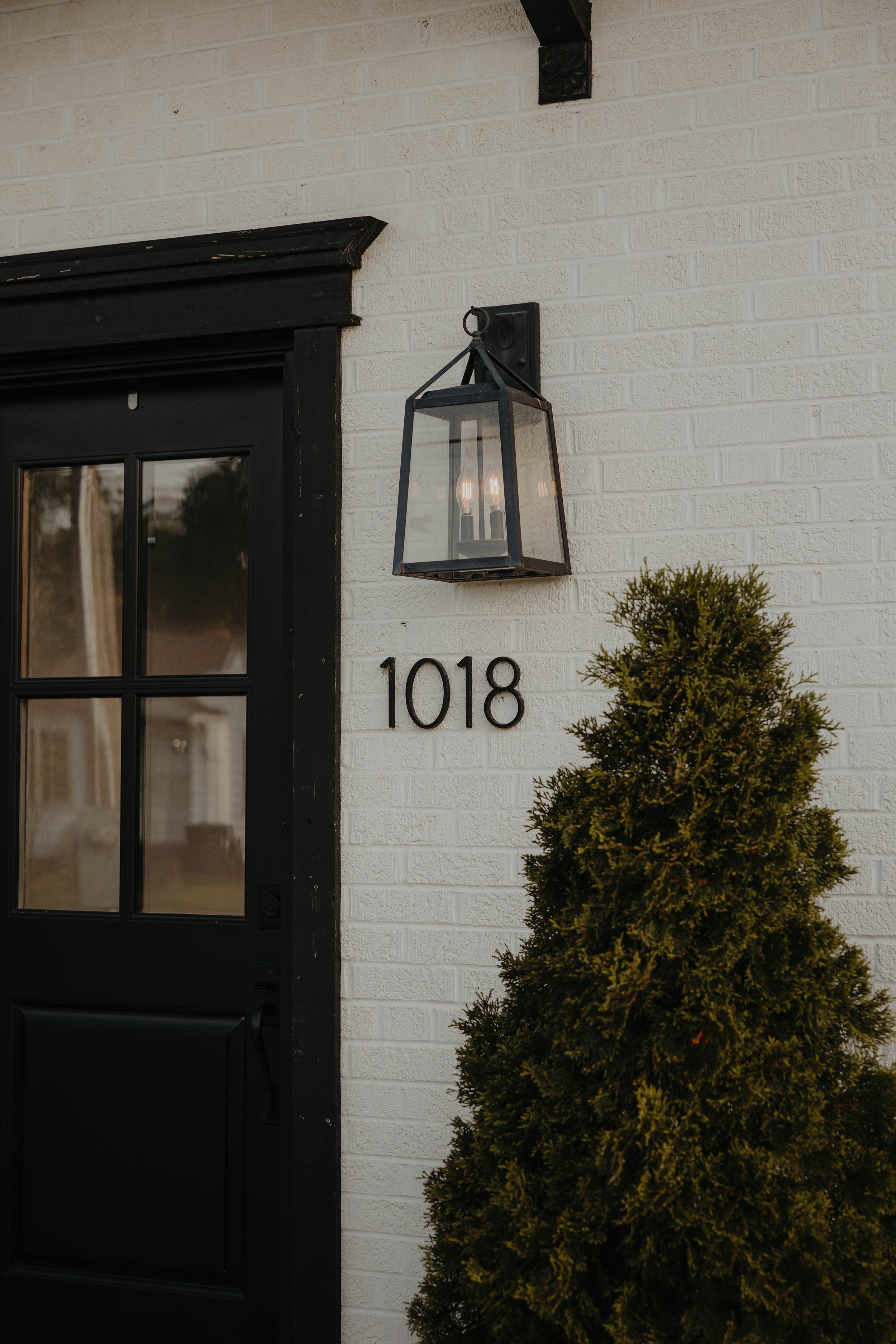 The image shows a white brick wall with the building number 1018 displayed on it. There is a black-framed glass door to the left, a black outdoor lantern mounted on the wall, and a green shrub in the lower right corner.