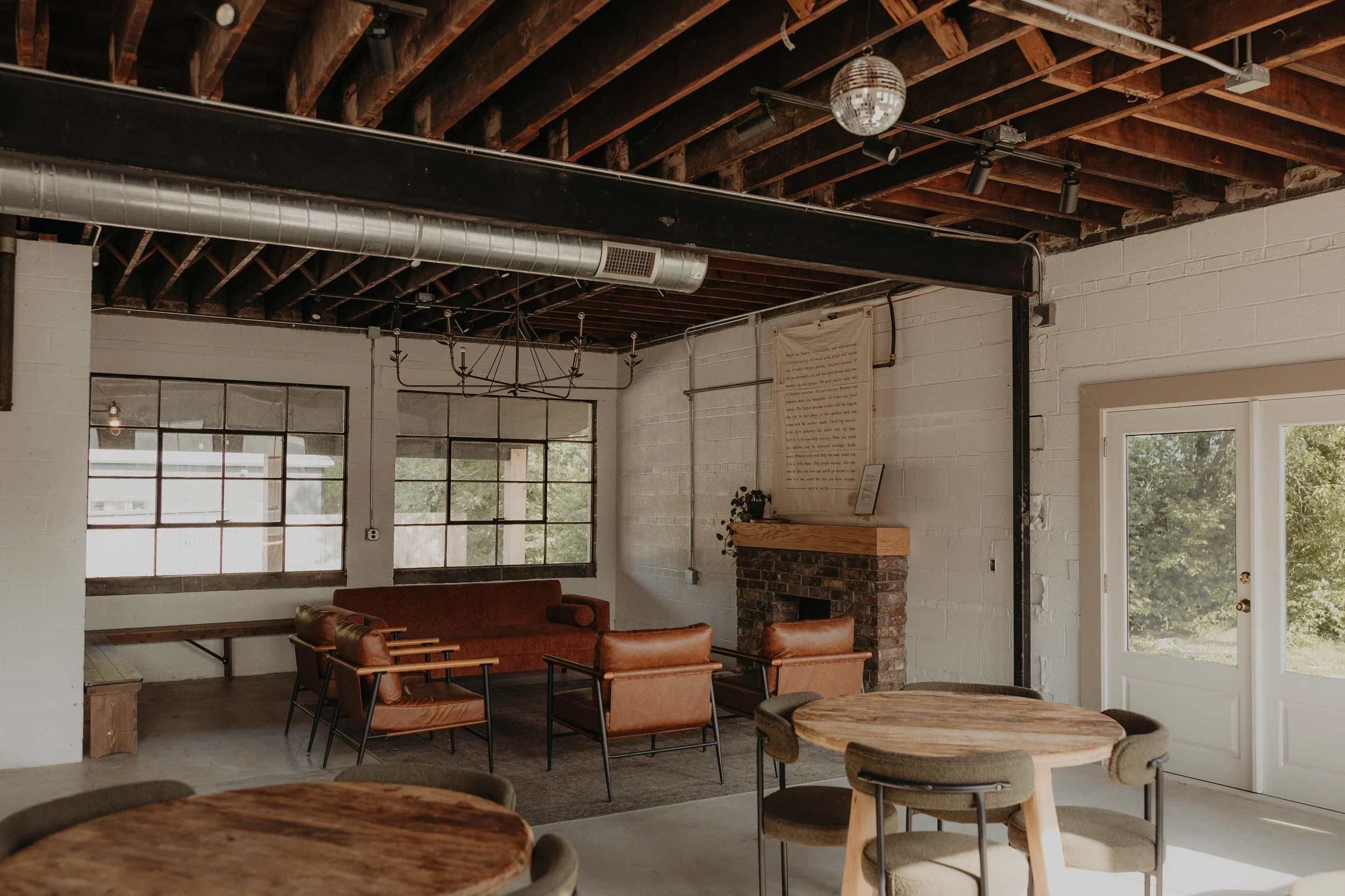 Interior of a modern industrial-style living space with exposed wooden ceiling beams, large windows, a brick fireplace, leather chairs, and wooden tables.