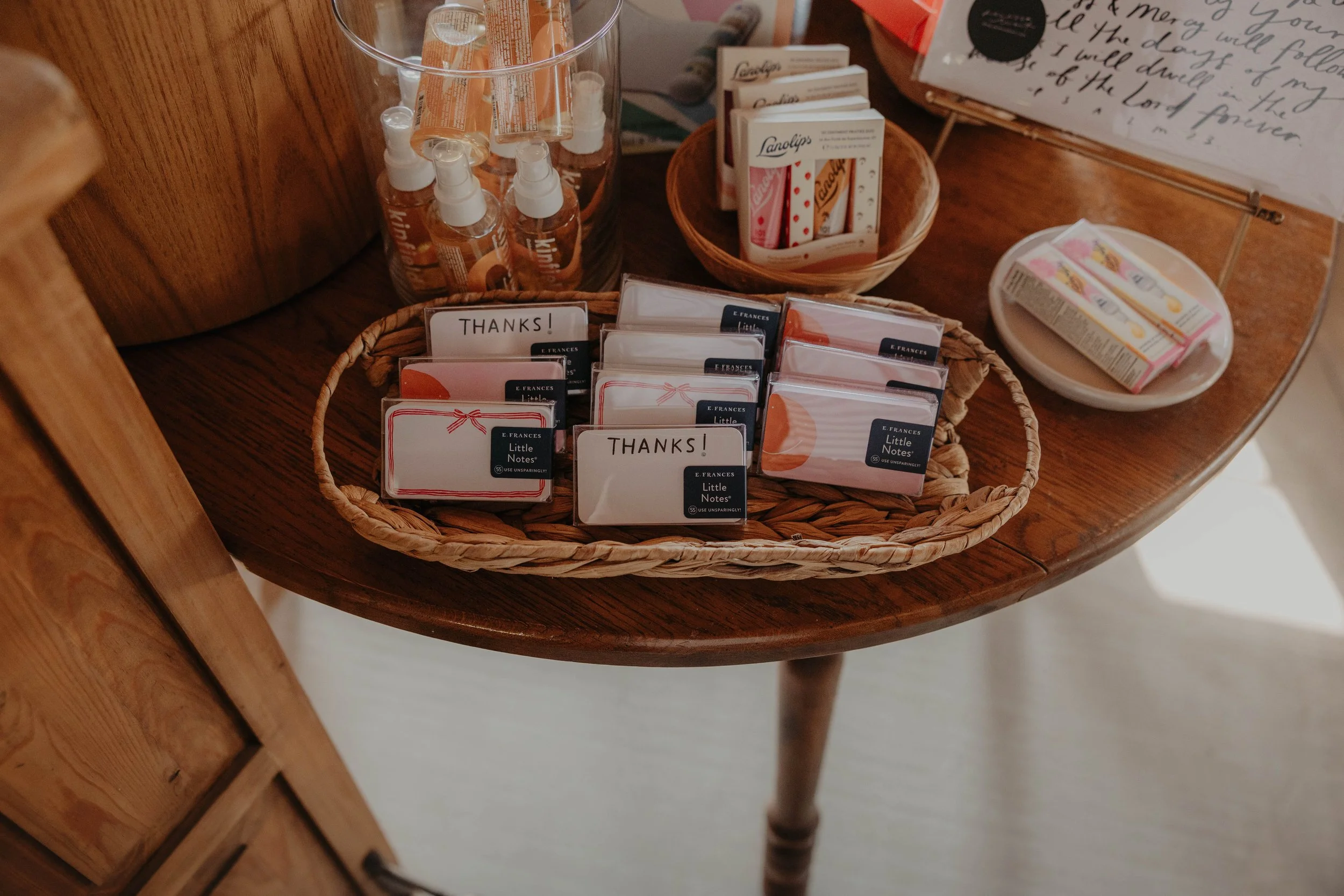 A wicker basket on a wooden table filled with small cards labeled 'Thanks!' and 'Little Notes,' and a clear container of small spray bottles of hand sanitizer. A small tray with a wrapped item and a artistic handwritten sign are also on the table.
