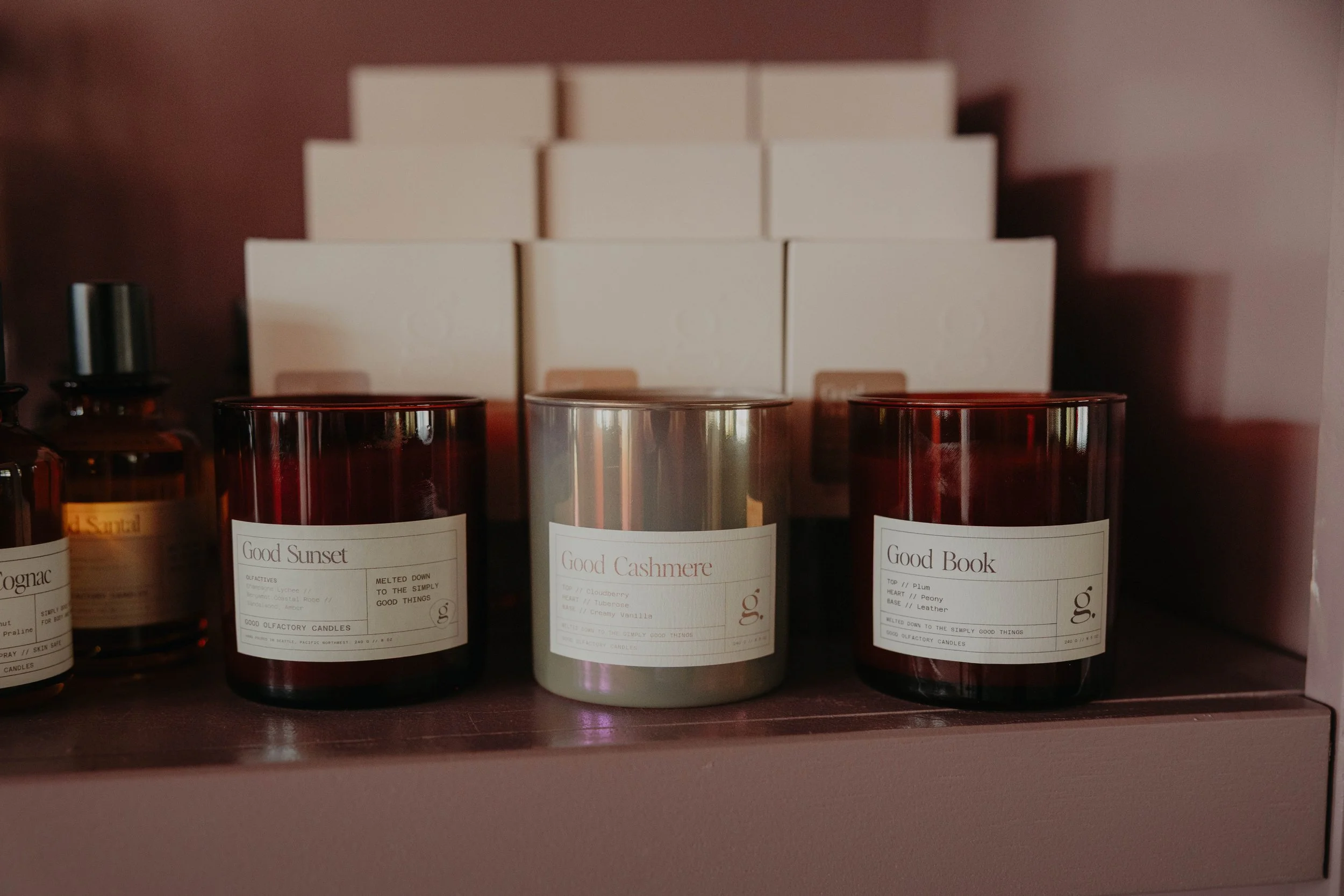Shelf with three candles labeled 'Good Sunset,' 'Good Cashmere,' and 'Good Book,' with boxes in the background.