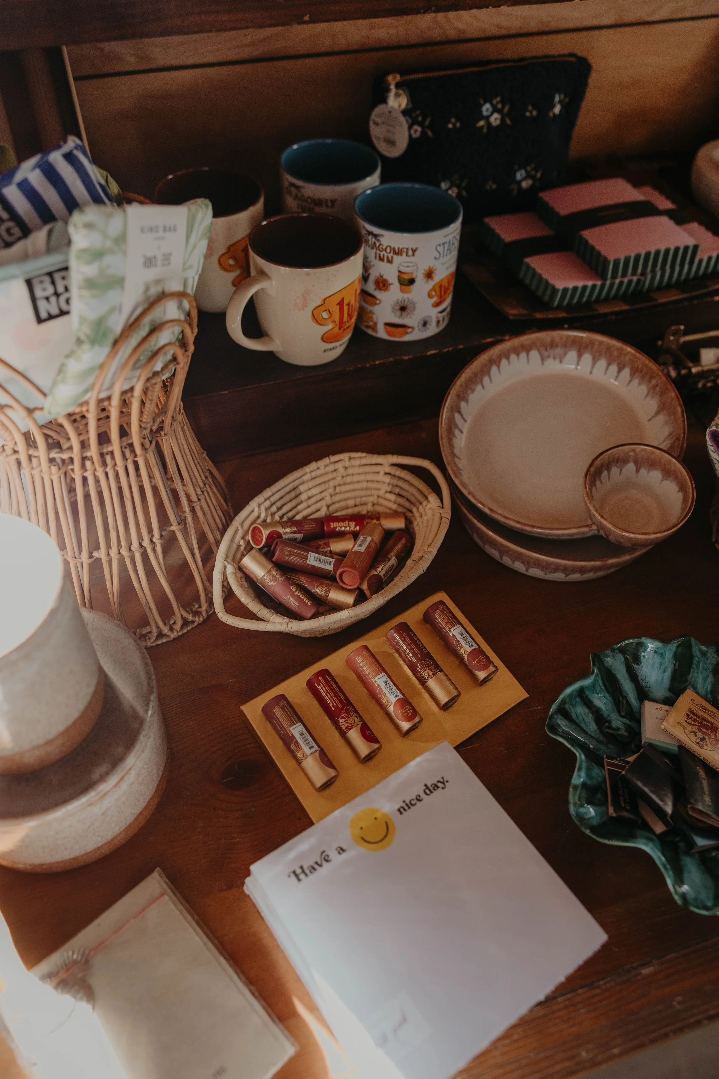 A display table with various items including ceramic mugs, small boxes, a woven basket with lip balm tubes, a ceramic bowl and smaller bowl, a black pouch, and stacks of pink and black striped napkins.