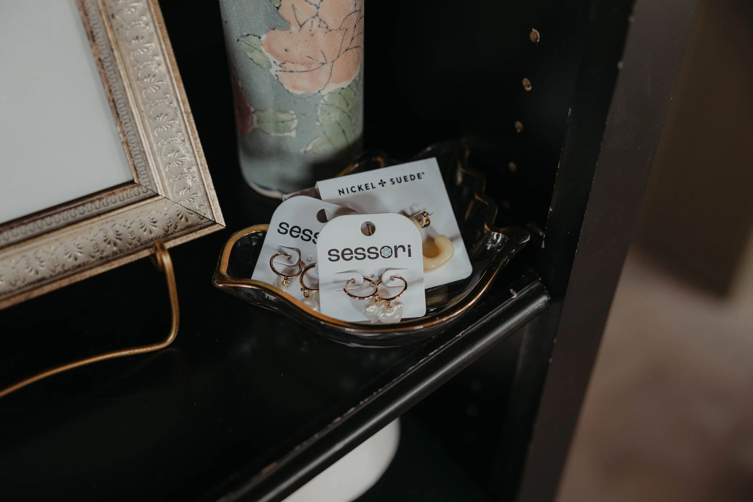 A black display shelf with jewelry, including gold hoop earrings with pearl accents, labeled 'sessori'. A ring and a beige-colored hair clip are also visible. Behind the jewelry, there is a decorative metal frame and a cylindrical container with a fl