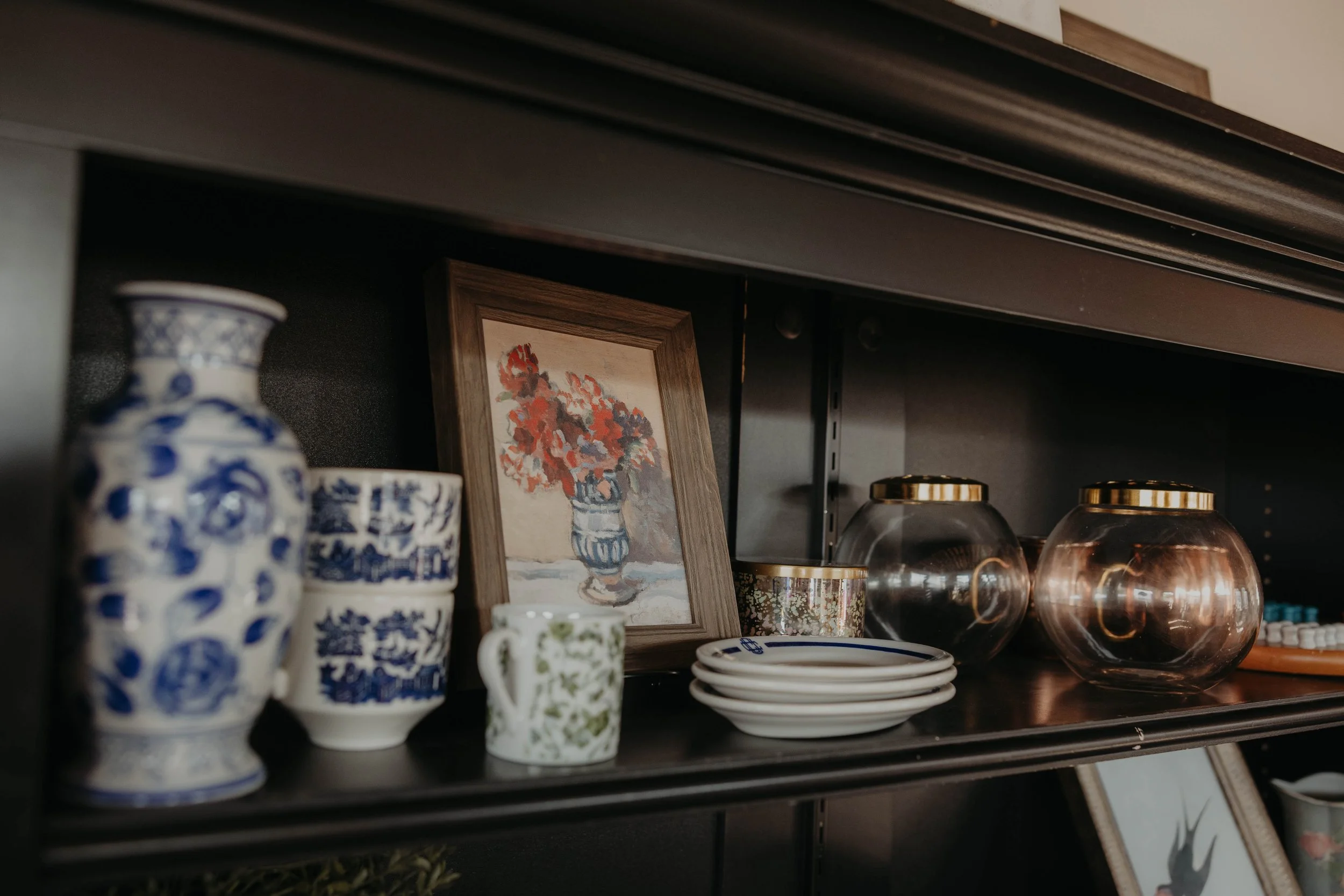 Shelf displaying blue and white patterned vases, small cups, plates, and glassware, with a framed floral artwork and decorative items behind them.