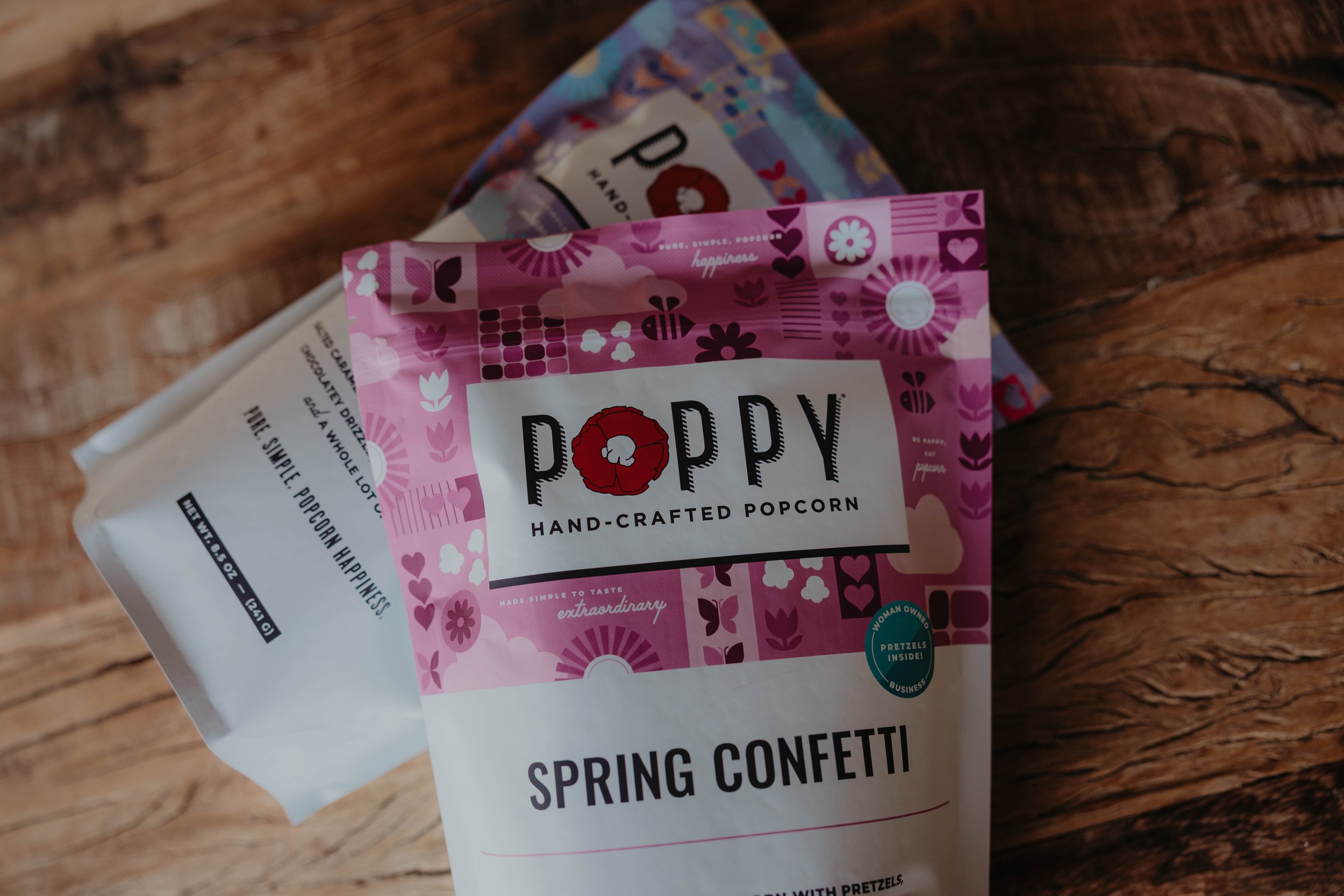 Pink popcorn bag with floral and heart patterns, labeled "Poppy Hand-Crafted Popcorn," resting on a wooden surface.