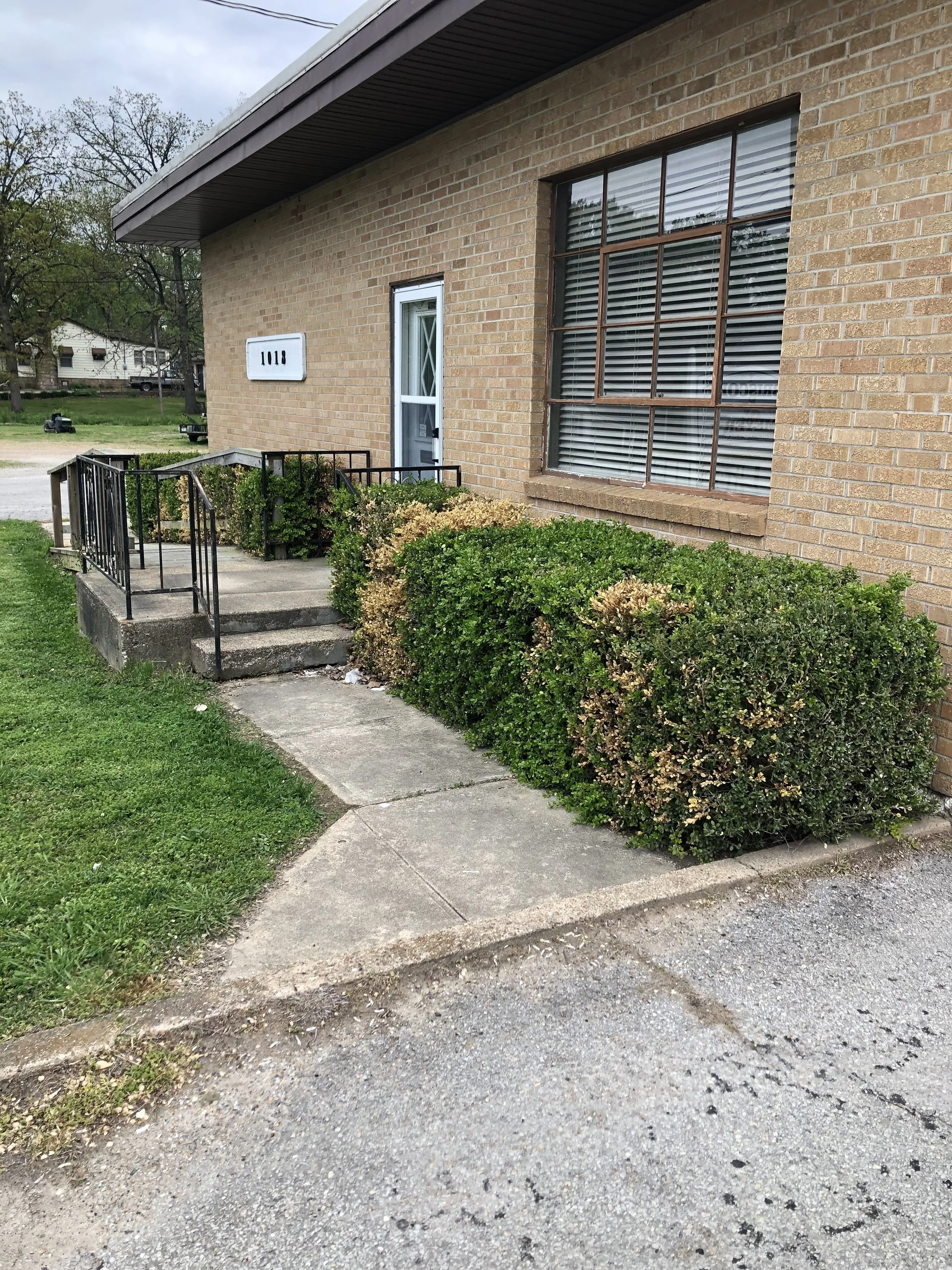 The photo shows the front of a brick building with large windows and a door. There's a small set of concrete stairs with a black metal railing leading up to the entrance. Green bushes and grass surround the sidewalk area, and a tree and other houses are visible in the background.