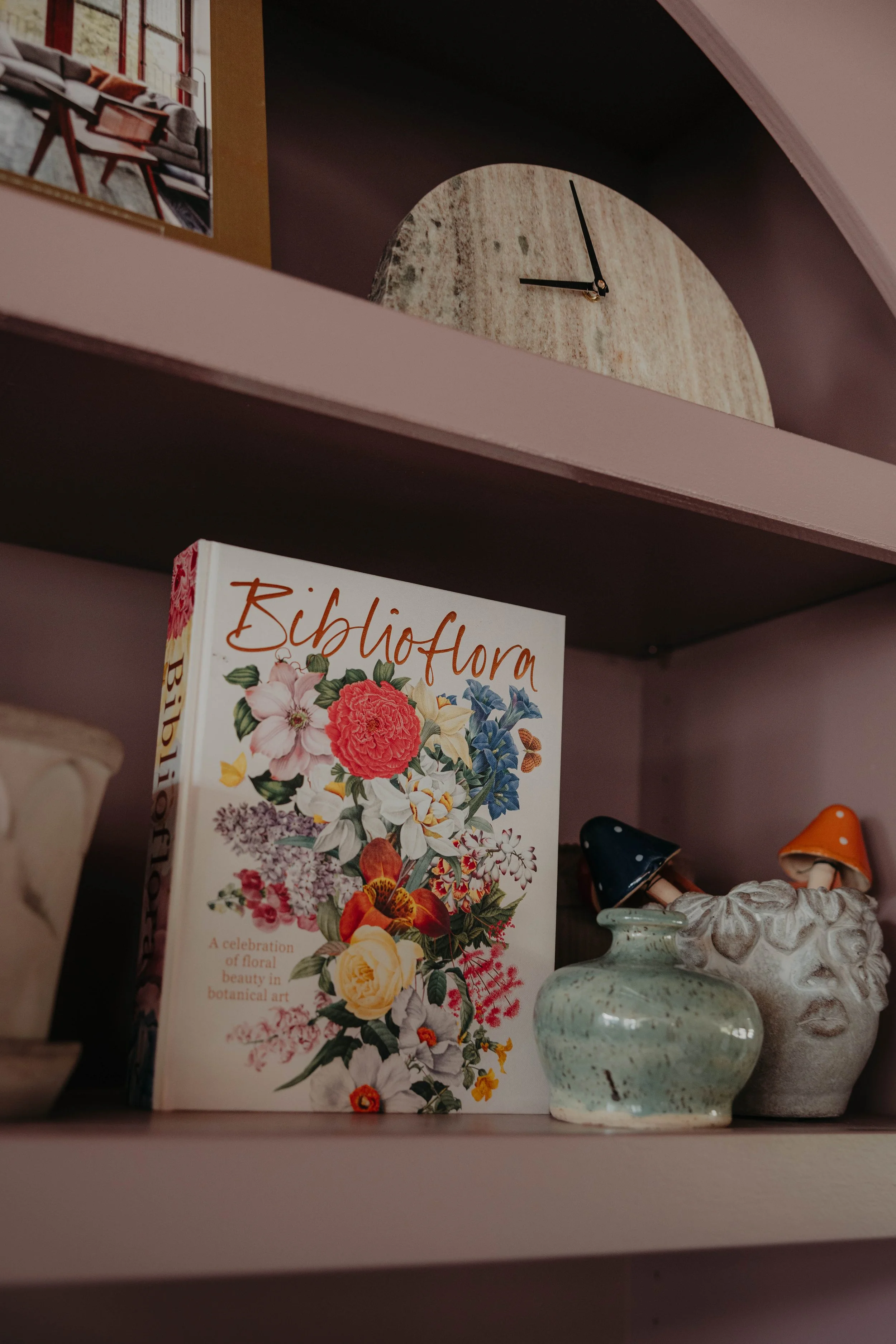 Close-up of a decorative floral book titled 'Biblioflora' standing on a shelf, with ceramic vases and small decorative mushrooms nearby, and a wall clock showing approximately 11:03 in the background.