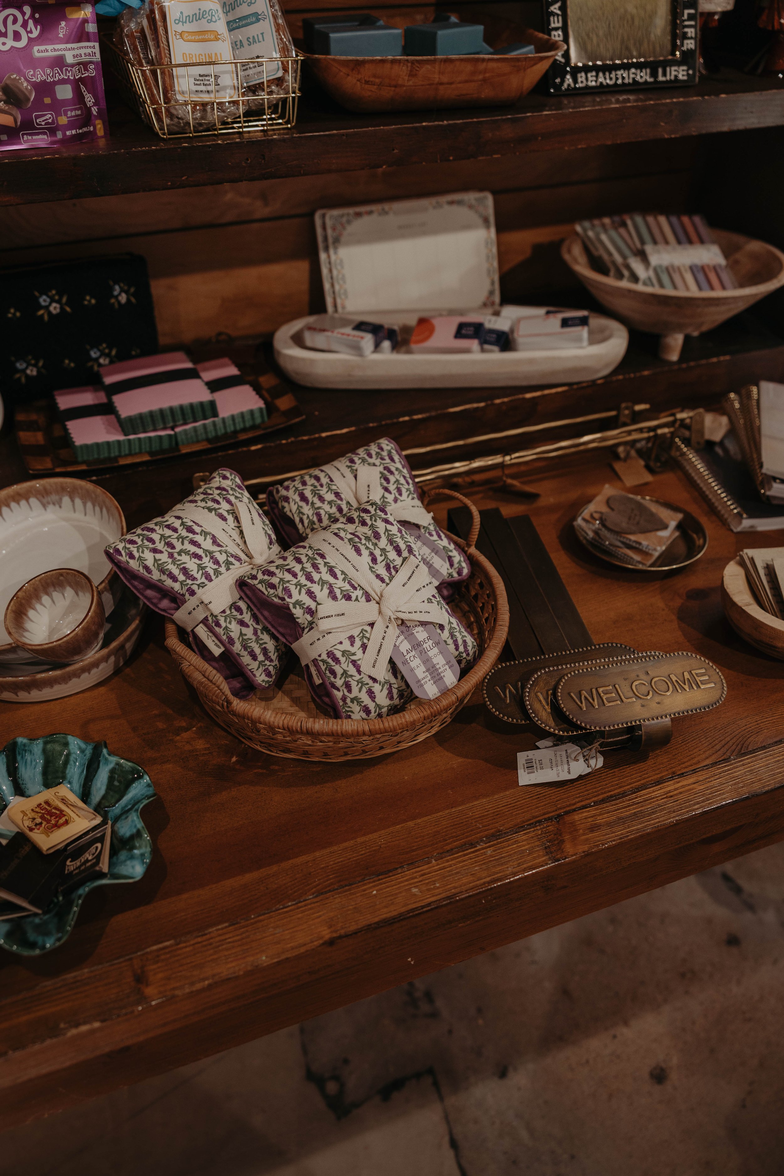 A wooden display table with various small home decor and gift items, including soap bars, a lavender pillow, small bowls, and decorative signs.