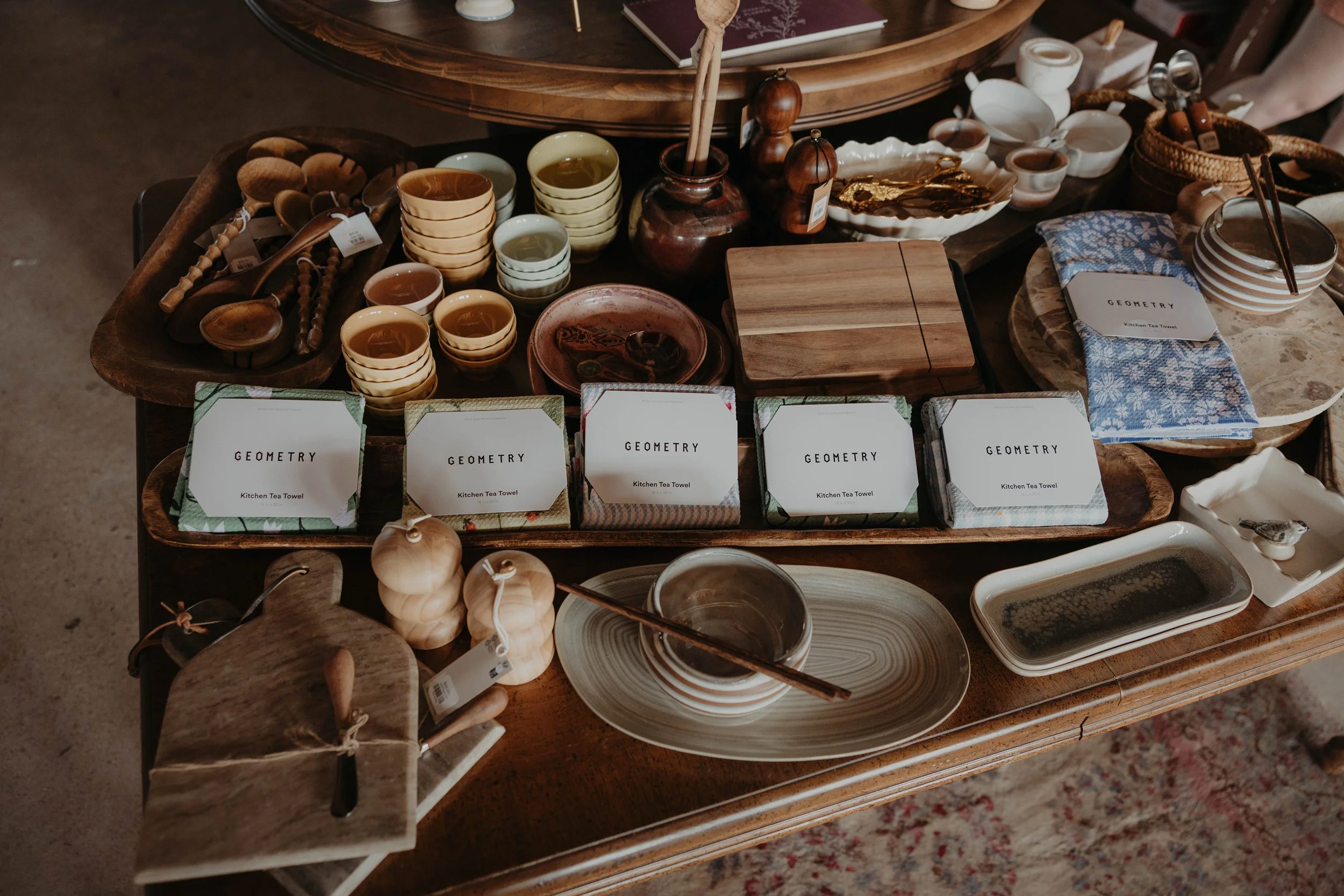 A wooden table displaying various ceramic and wooden kitchenware items, including cups, bowls, spoons, a chopping board, and decorative objects, with tags indicating they are from the brand 'GEOMETRY'.
