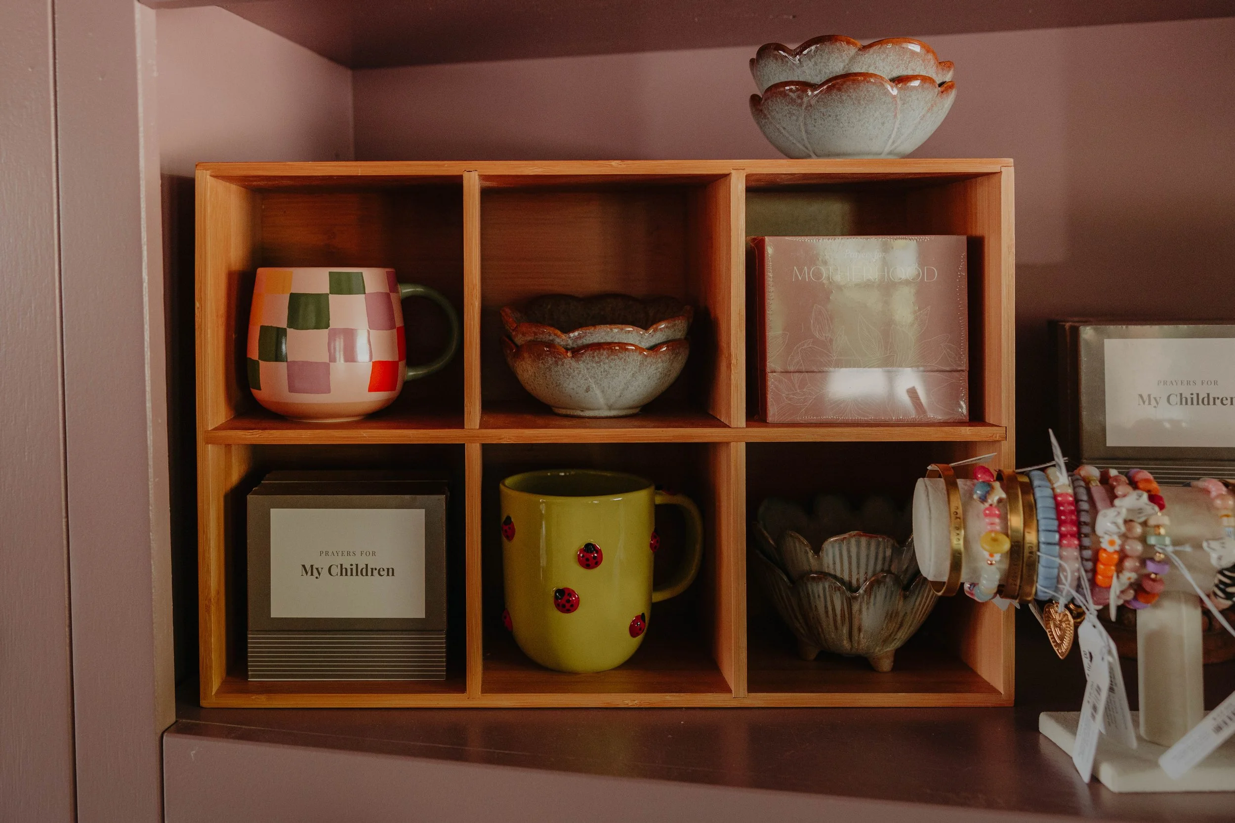 A wooden shelf with compartments holding colorful ceramic mugs, bowls, framed pictures, and jewelry displays.