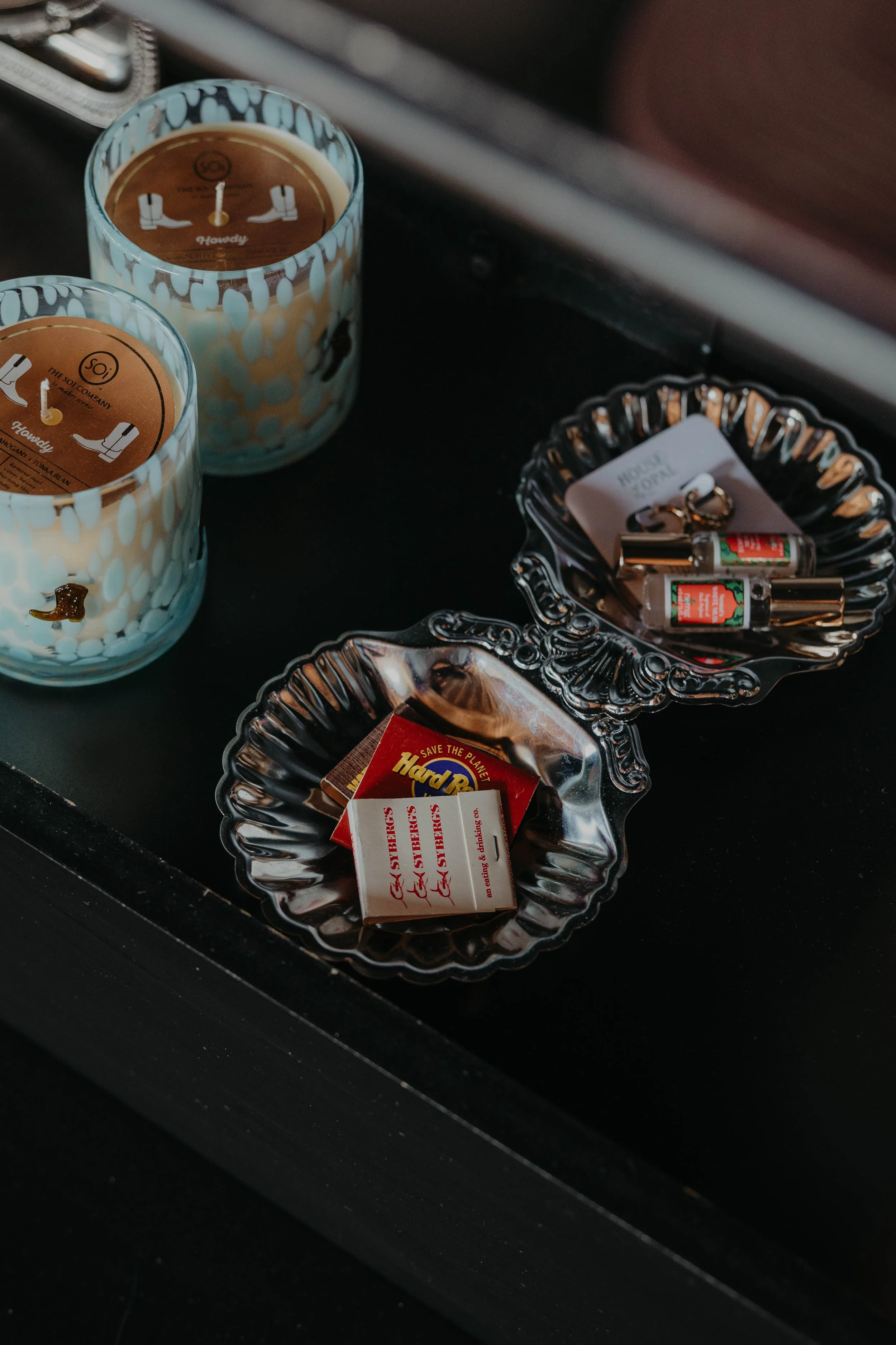 Two candles in glass holders with decorative patterns, and a silver tray containing small bottles, a matchbox, and small boxes on a black surface.
