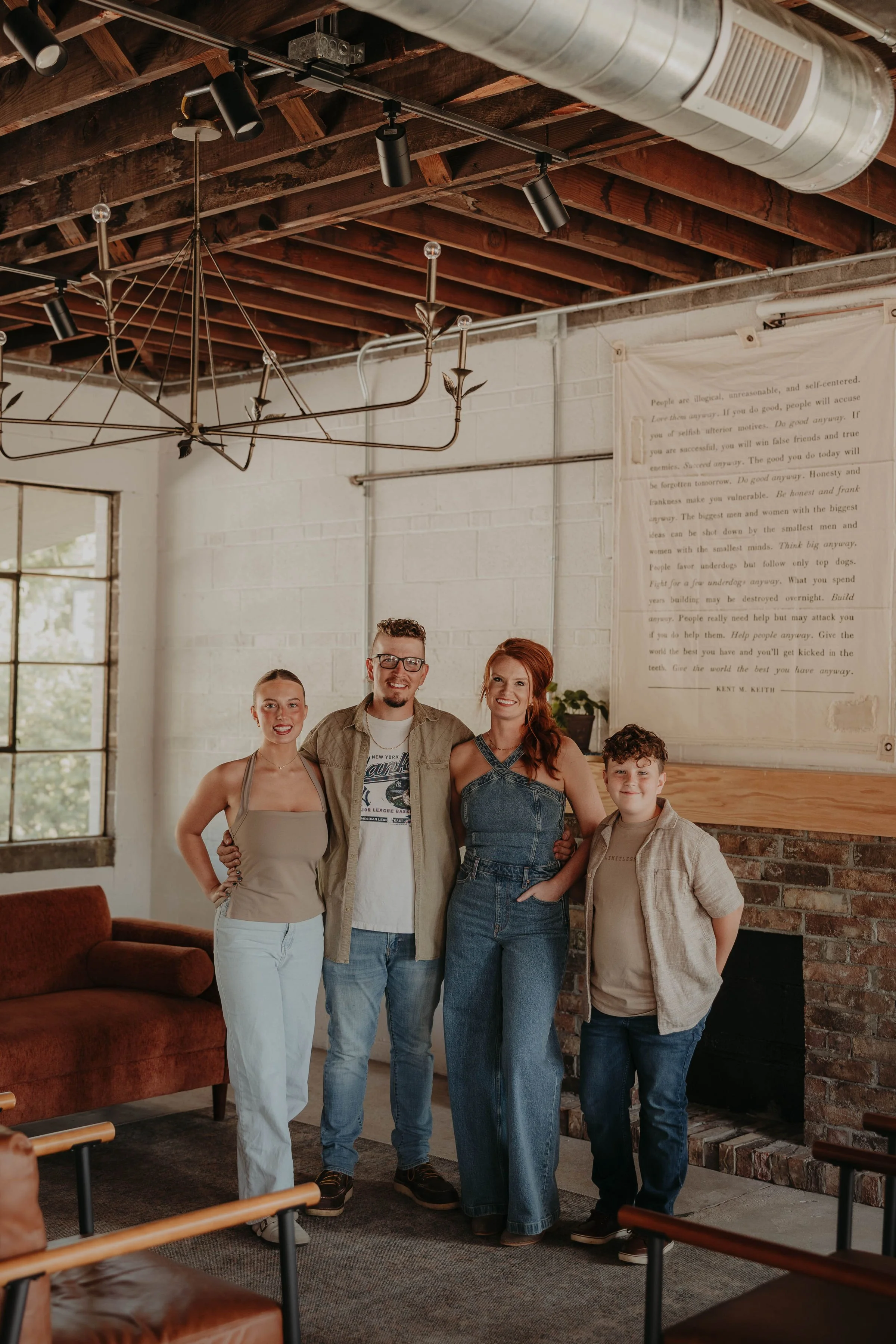 Family of 4 standing together in a modern industrial-style room with exposed brick walls and wooden beams. They are smiling and posing for the photo, with a large white text banner on the wall and a dark red sofa partially visible in the foreground.
