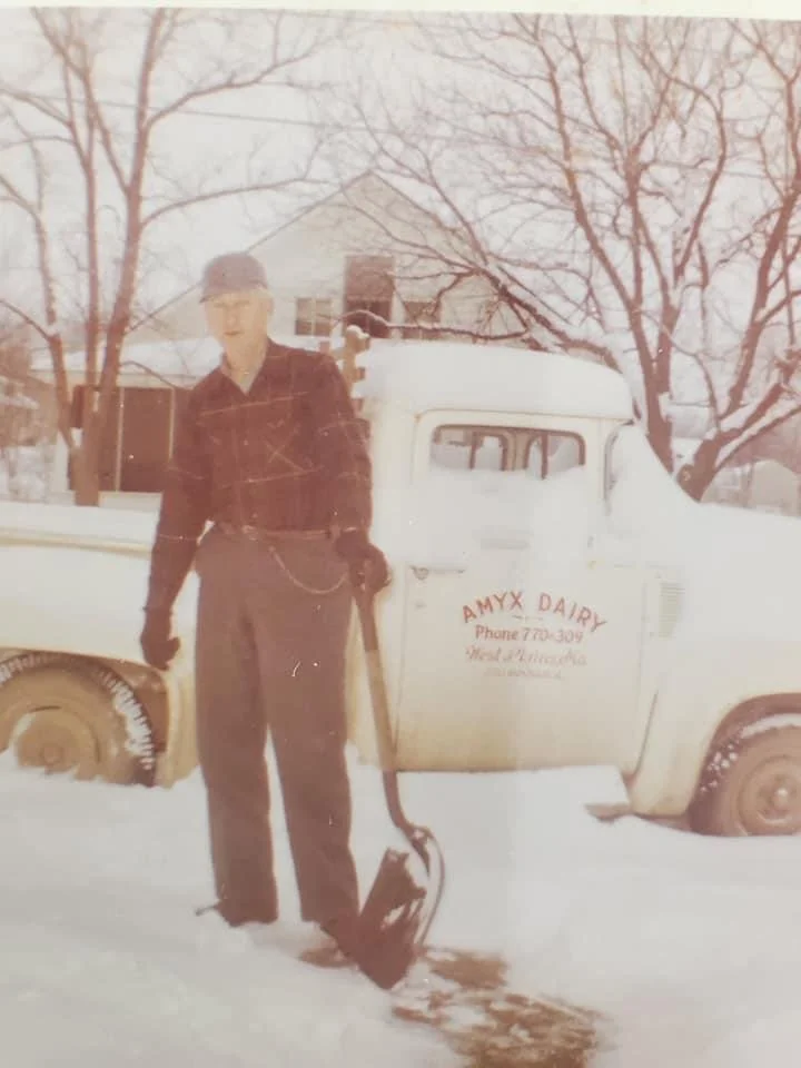A man dressed in vintage clothing, standing in snow next to a classic pickup truck with 'AMYX DAIRY' written on the door, holding a shovel with snow on it.