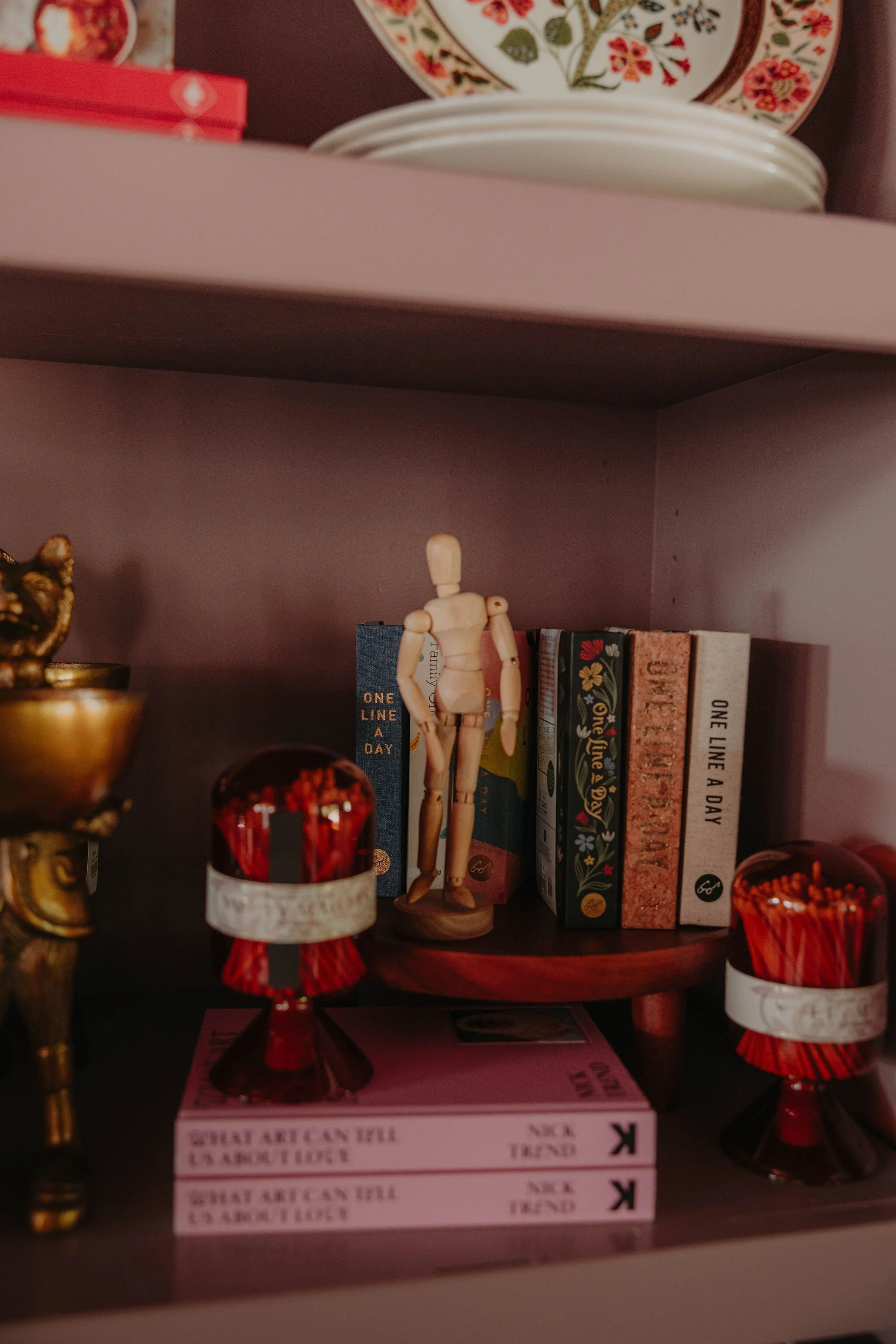 A bookshelf with decorative plates on the top, a small wooden mannequin figure, and books including titles like "Great Expectations" and "One Line a Day," with some candle jars or small decorative bottles in the foreground.