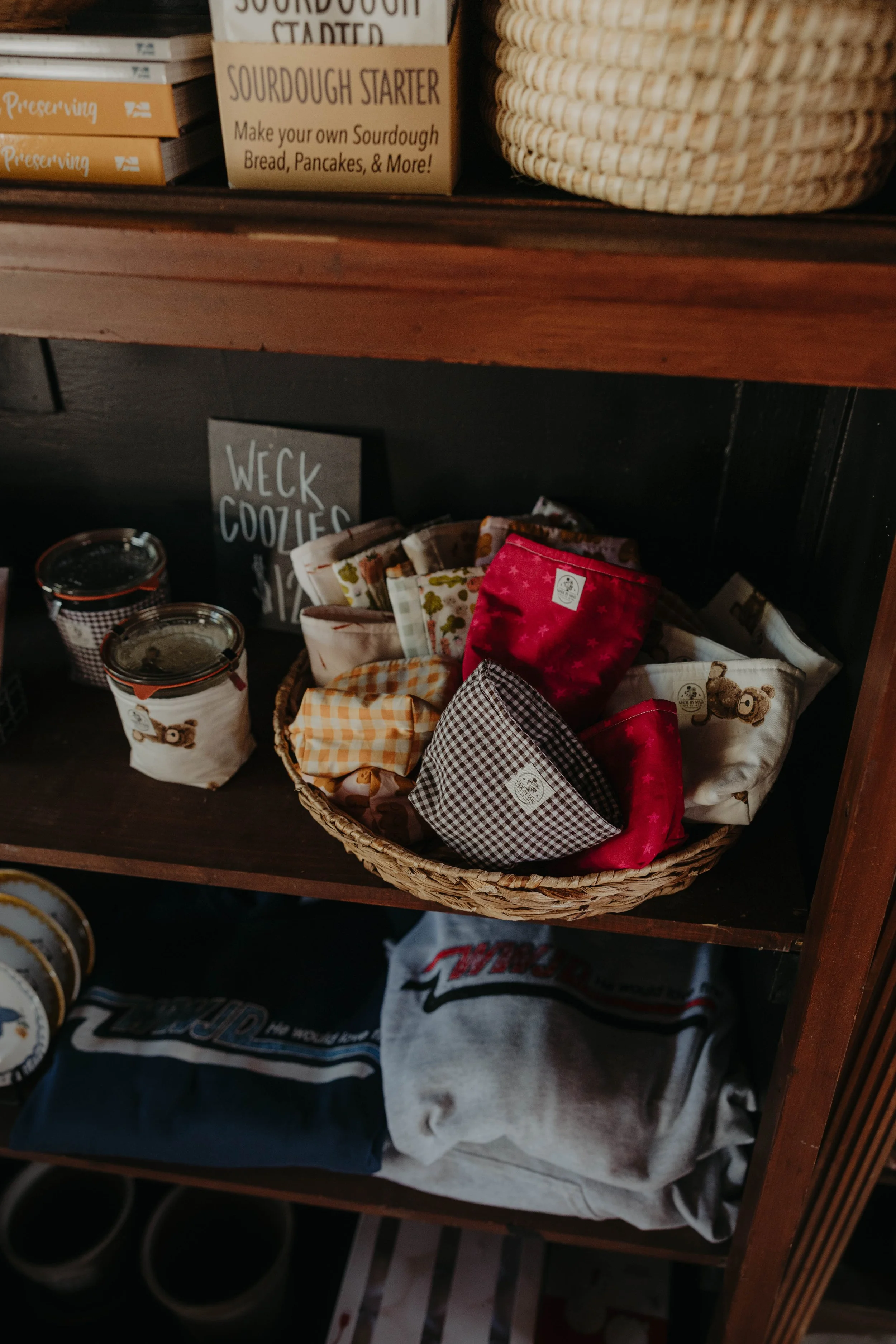 A wooden shelf displays various items including a basket of fabric items, a sign with the word 'WECK COOKIES,' tin cans, and boxes labeled 'Sourdough Starter' and 'Preserving.'