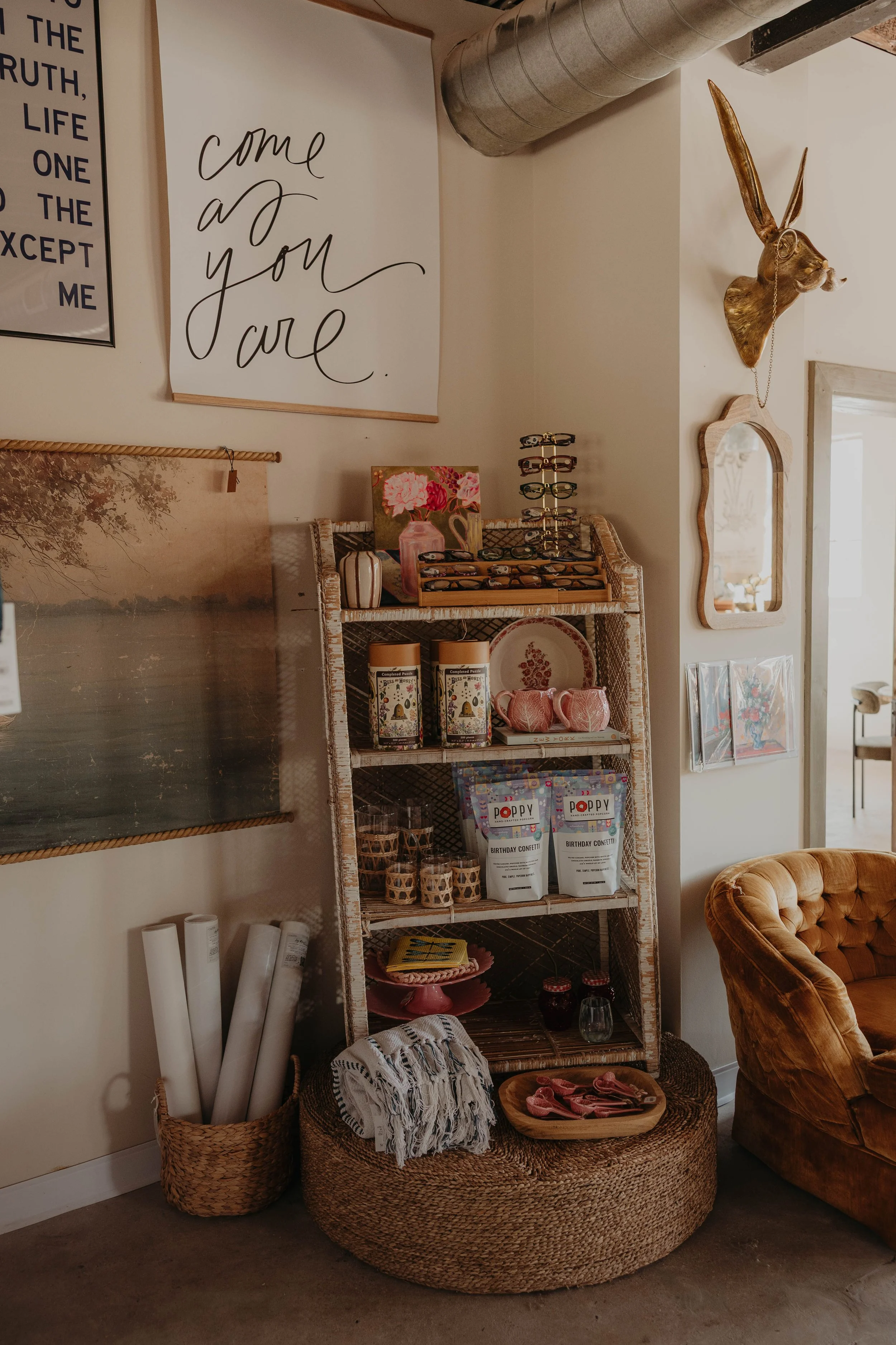 Image of a cozy corner in a home with a wicker shelving unit, botanical wall art, a mounted deer head with glasses, framed pictures, and various decorative items including pottery, candles, and gift boxes. There's a plush orange armchair nearby and w