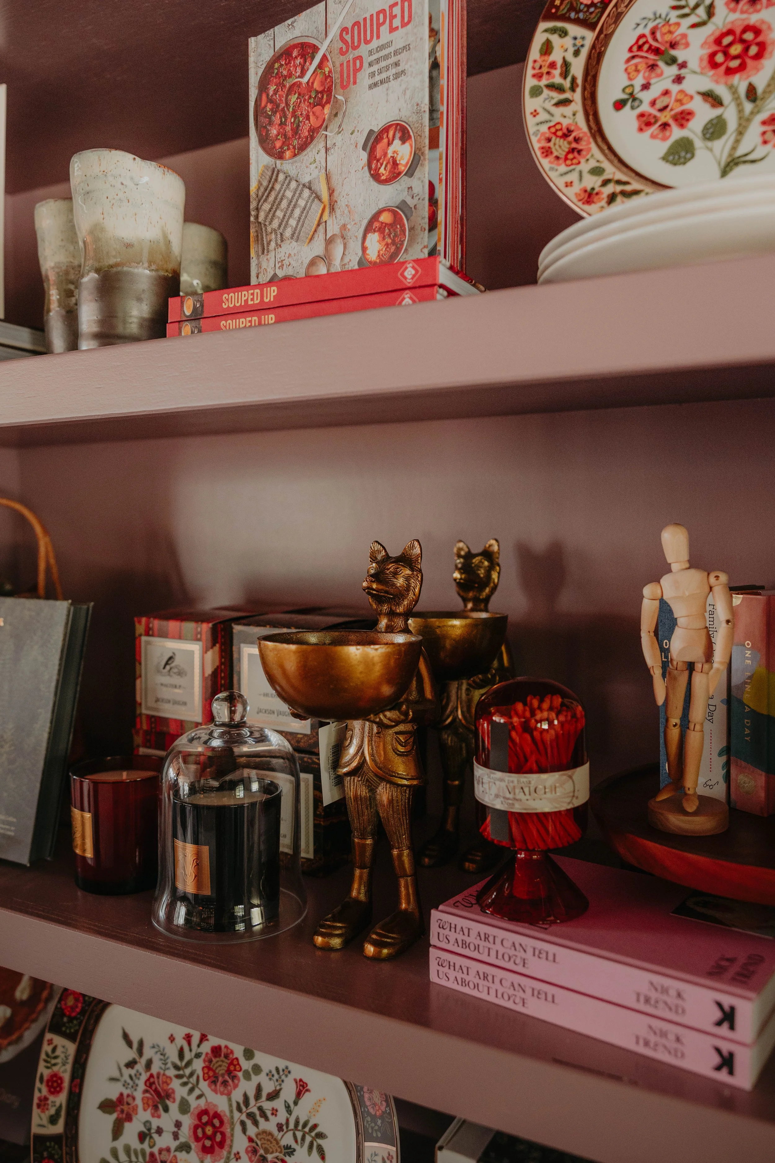 A pink shelf displaying various decorative and functional items, including a gold Scottish-themed goblet, a glass dome, candles, a figure of a person, and colorful plates with floral designs. There are also books and boxes with fashionable and artist