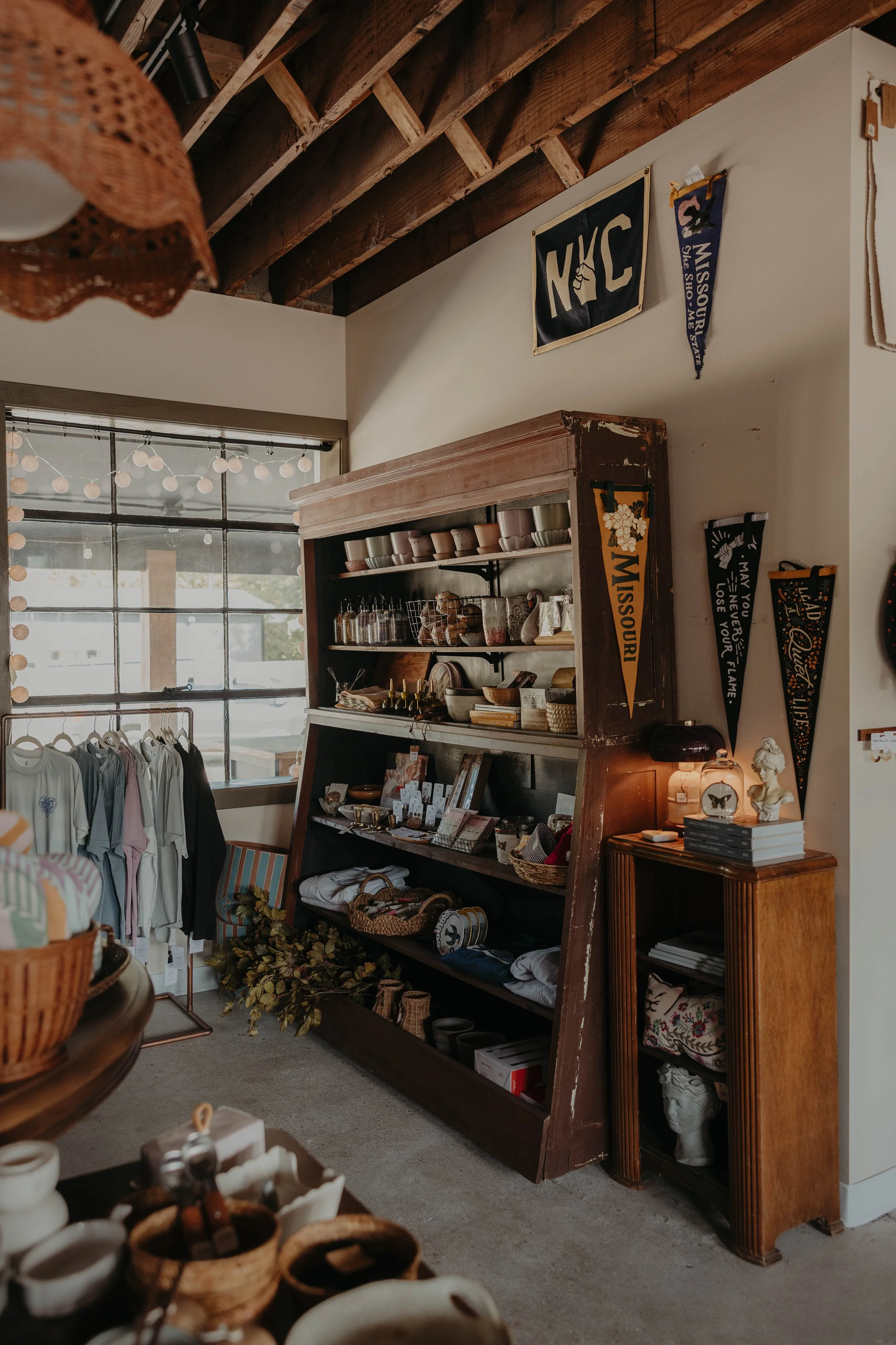 Interior of a vintage-style boutique store with wooden ceiling beams, a large window, and various home decor items, clothing, and accessories on display.