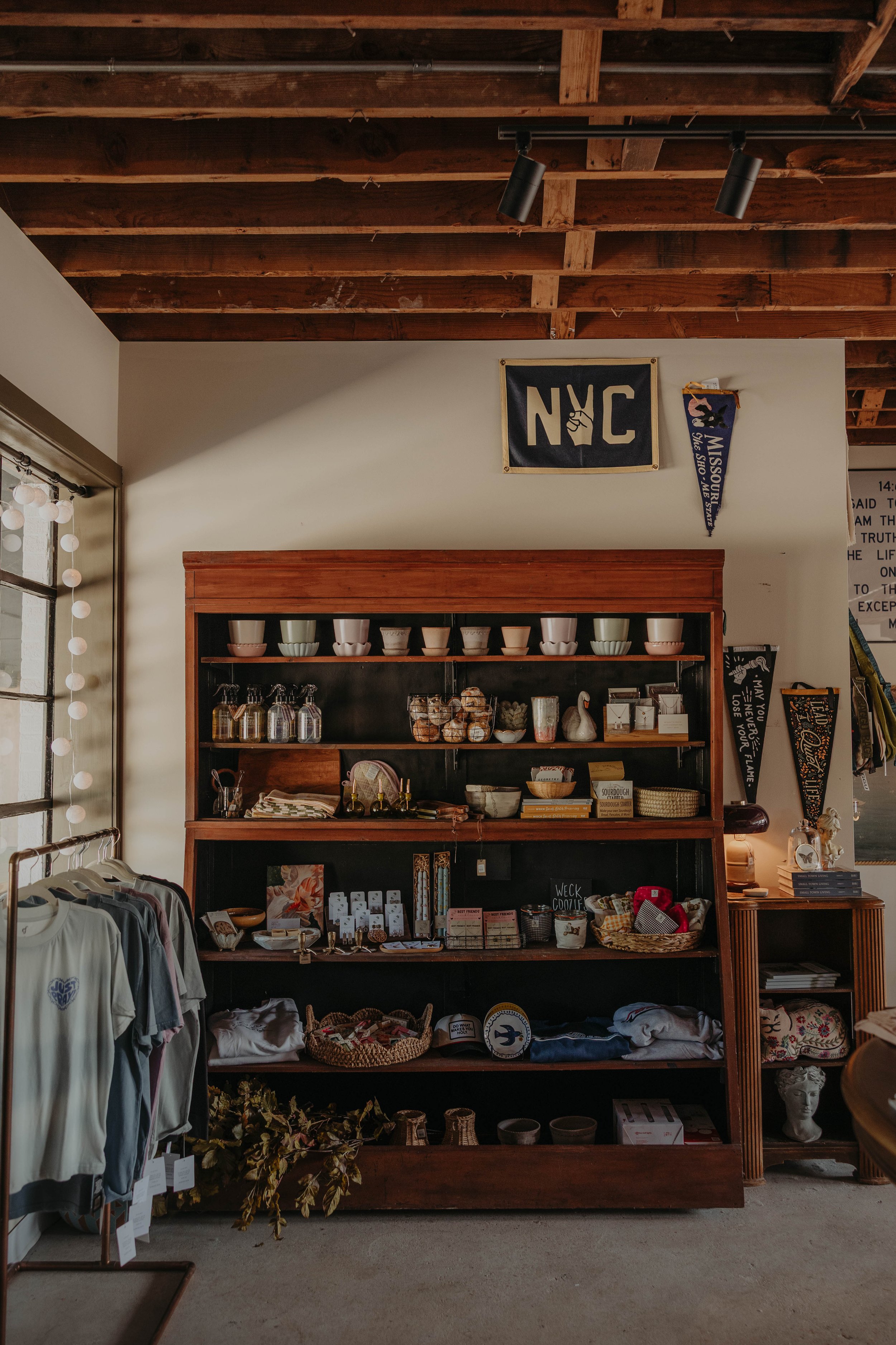 Interior of a store with wooden shelves displaying bowls, jewelry, and small decorative items. To the left, clothing items are hanging on a rack. There are banners and signs on the wall, including one with 