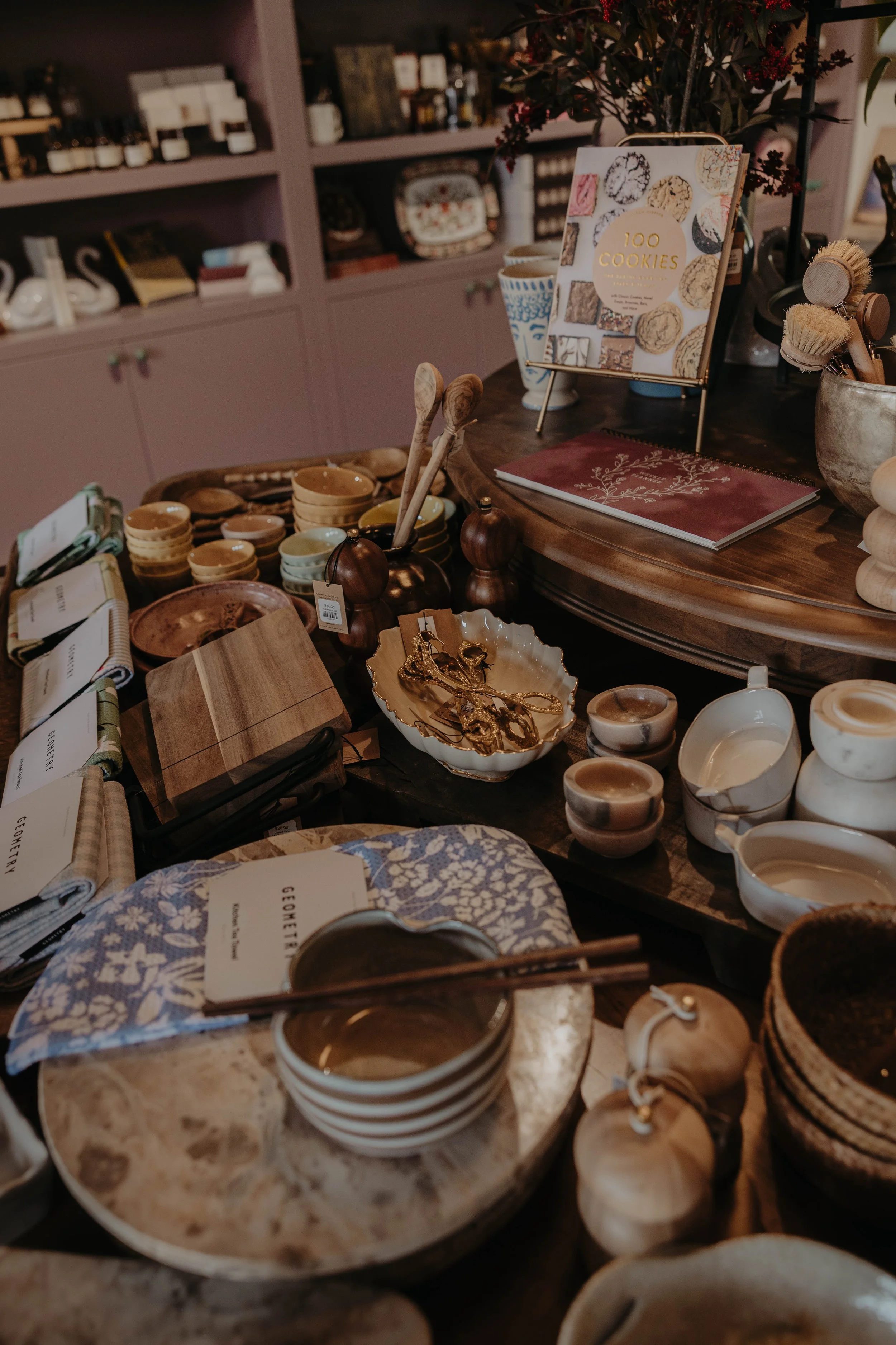 A display of various kitchenware items on a wooden table, including bowls, utensils, and sprigs of herbs, with a book titled '100 Cookies' and a set of decorative brushes nearby.