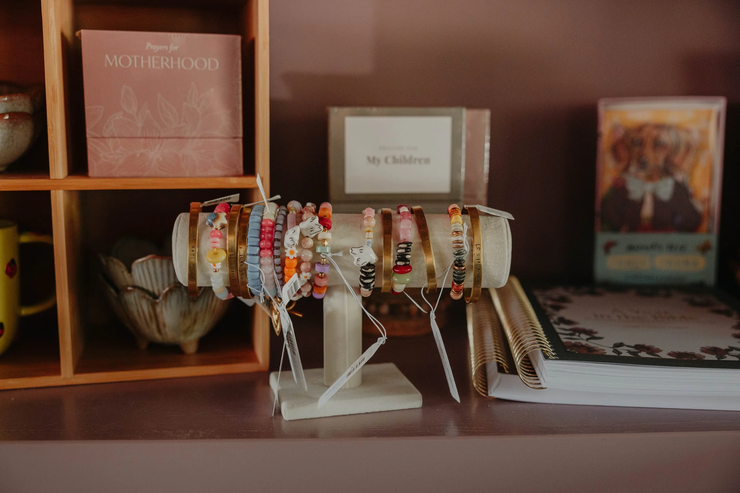 Display of assorted colorful beaded bracelets on a jewelry stand in front of wooden shelves with books and decorative items.