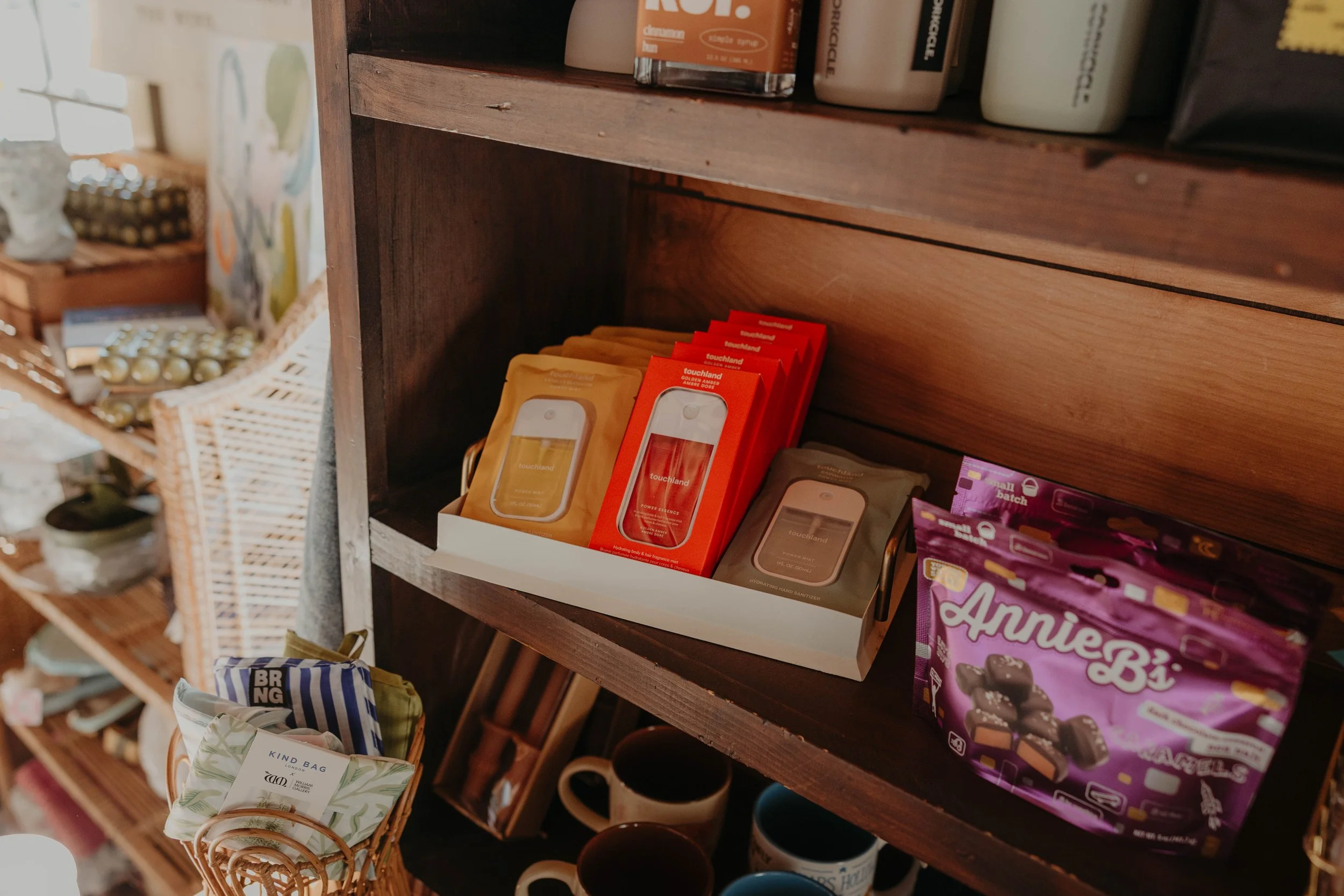 Wooden shelves displaying various products including hand sanitizer, chocolate, mugs, and snack bags in a retail store.