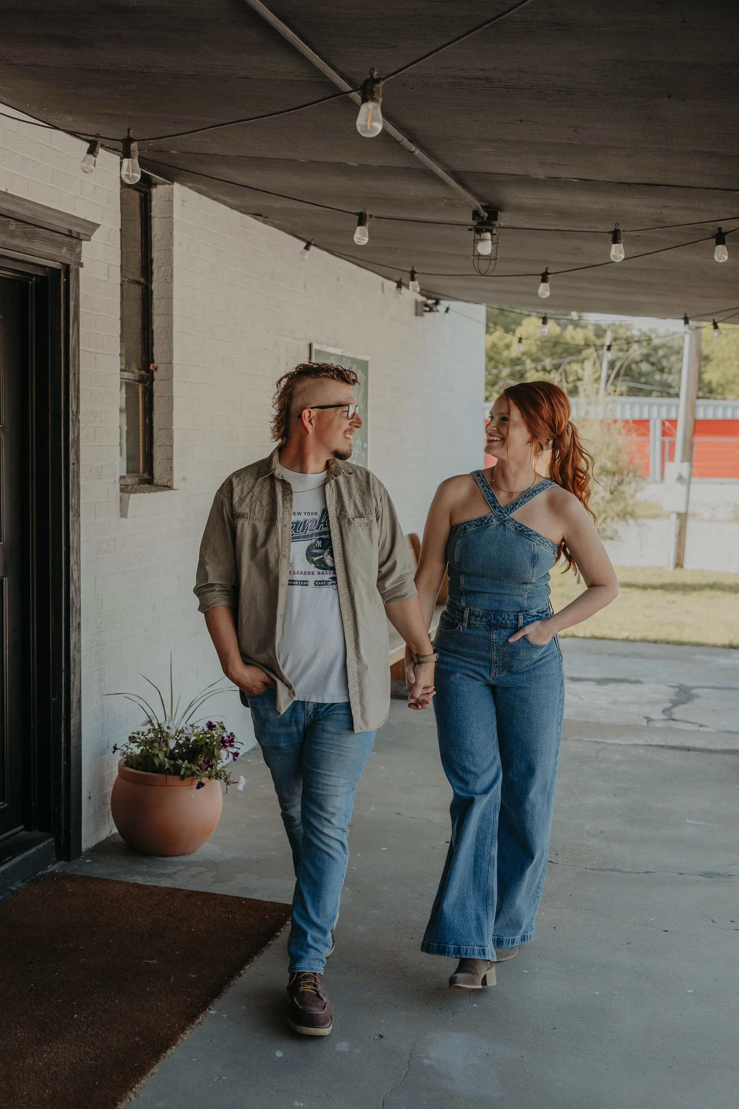A man and woman walking hand in hand under a covered outdoor area, smiling at each other. The woman has red hair and is wearing a denim jumper, while the man has glasses and curly hair, dressed in a casual jacket and jeans. There is a potted plant with flowers nearby, and string lights hanging from the ceiling.