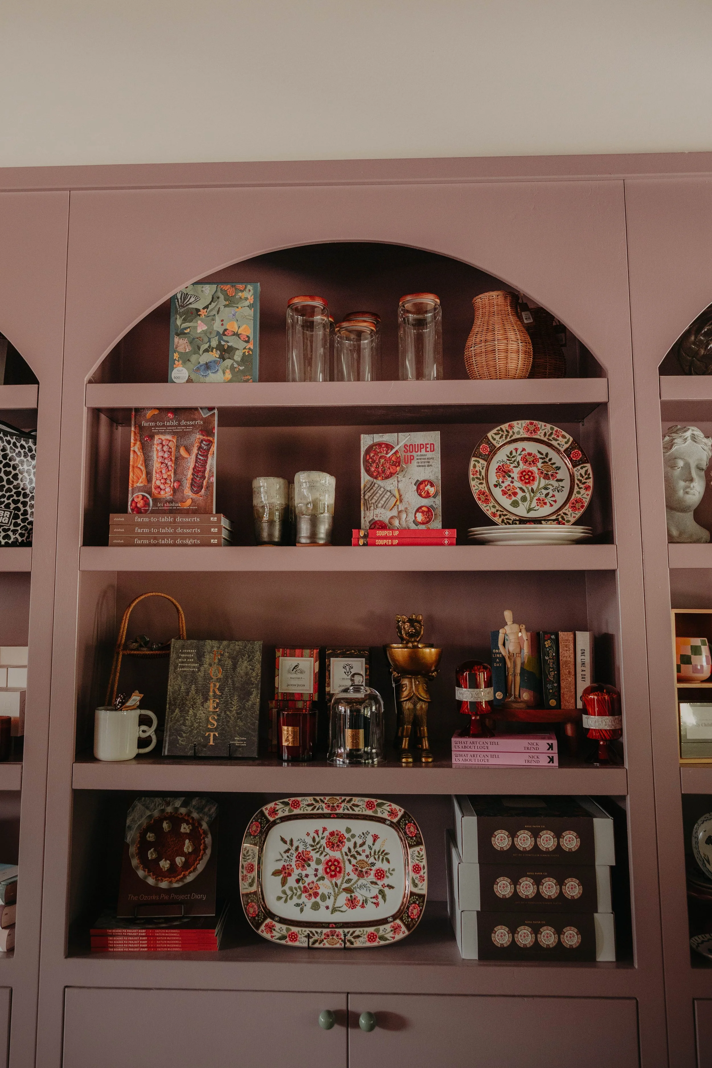 A pink cabinet with decorative dishes, glass jars, a wicker vase, books, and various decorative items on its shelves.