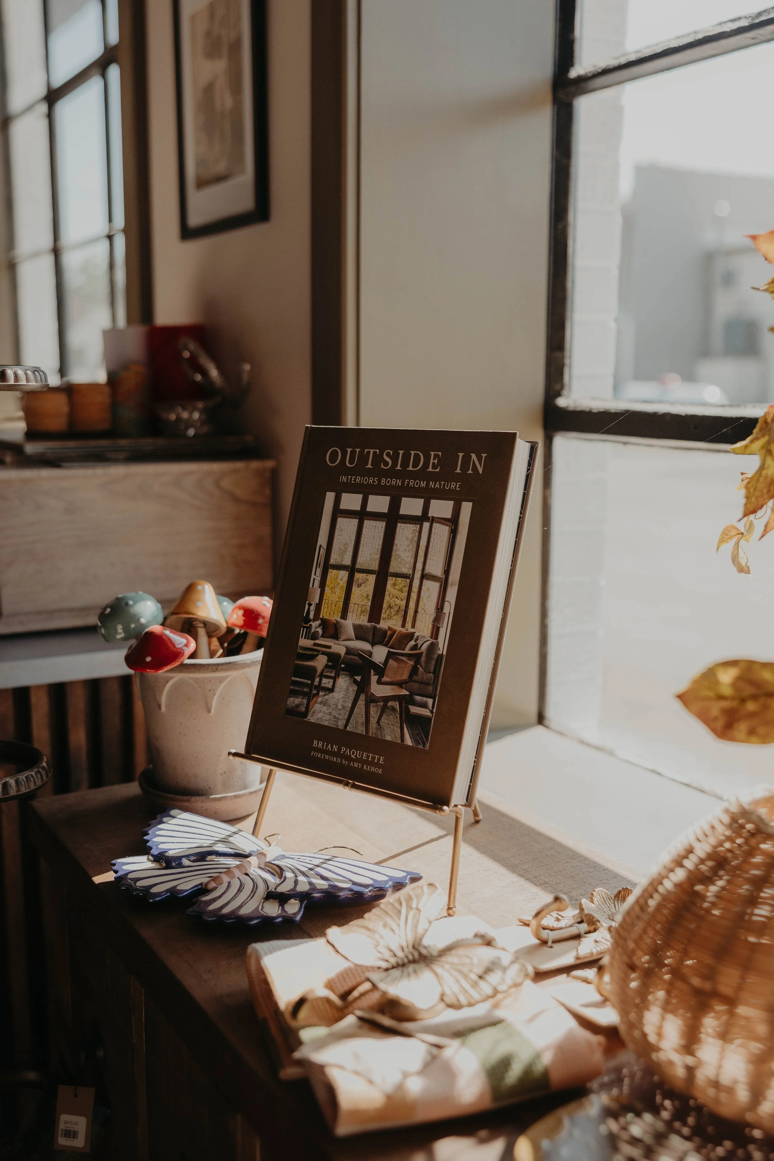 A display of various home decor items, including a book titled 'Outside In' by Brian Paquette, decorative mushrooms, shells, and woven baskets on a wooden table near a window with sunlight streaming in.