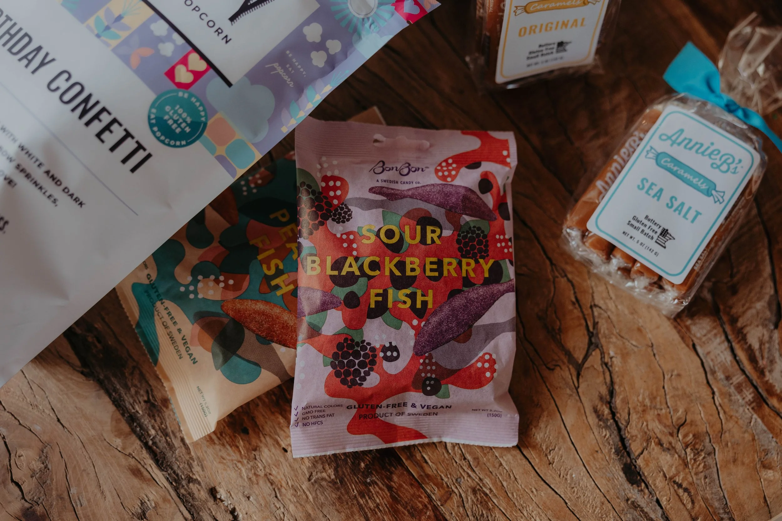 Various snack packages on a wooden surface, including a bag of sour blackberry fish, sea salt caramel candies, and other snack items.