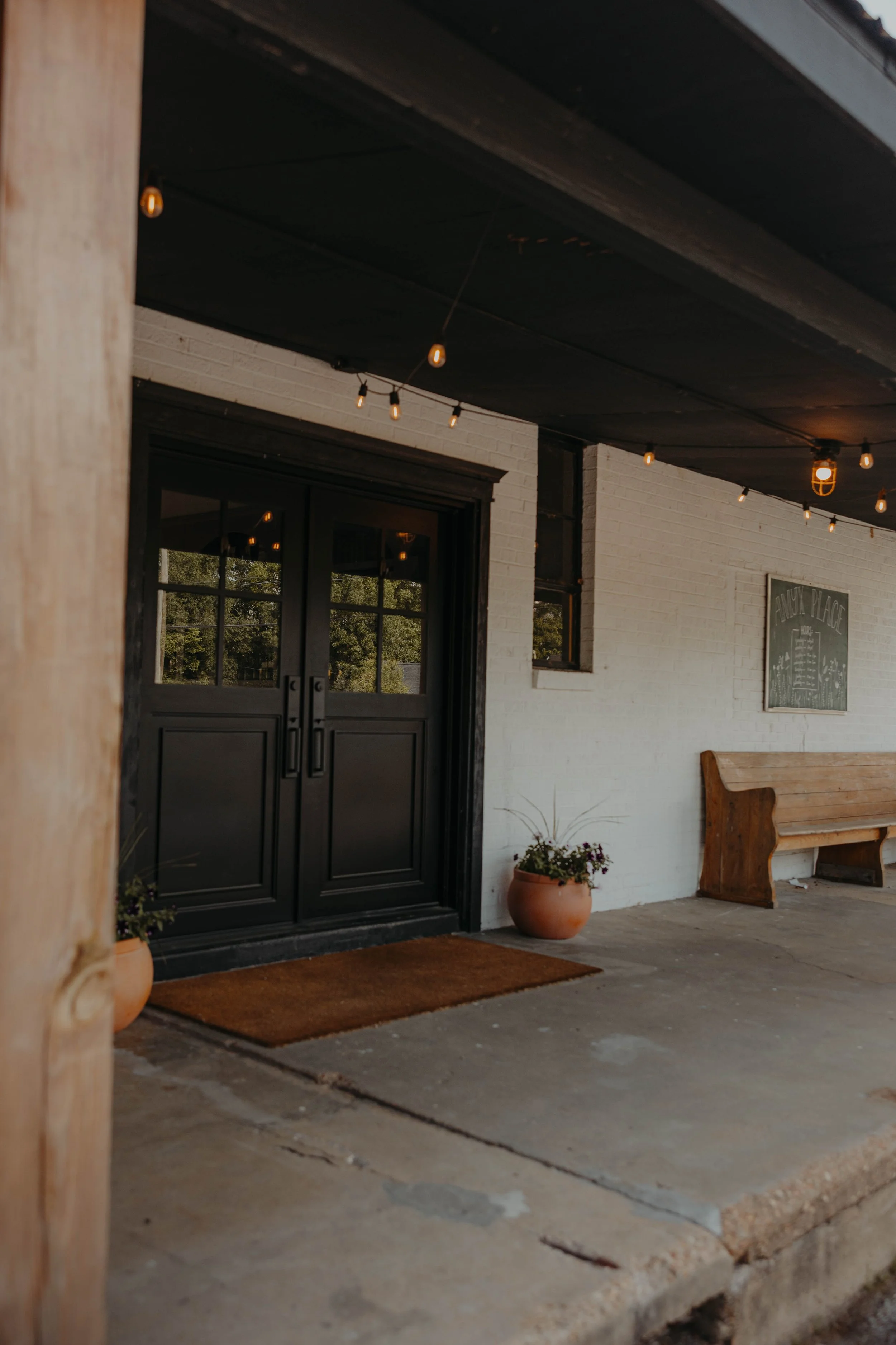 The image shows the entrance of a building with black double doors, a small window on the right, a white brick wall, string lights hanging from the dark ceiling, and potted plants outside. There is also a wooden bench and a chalkboard sign.