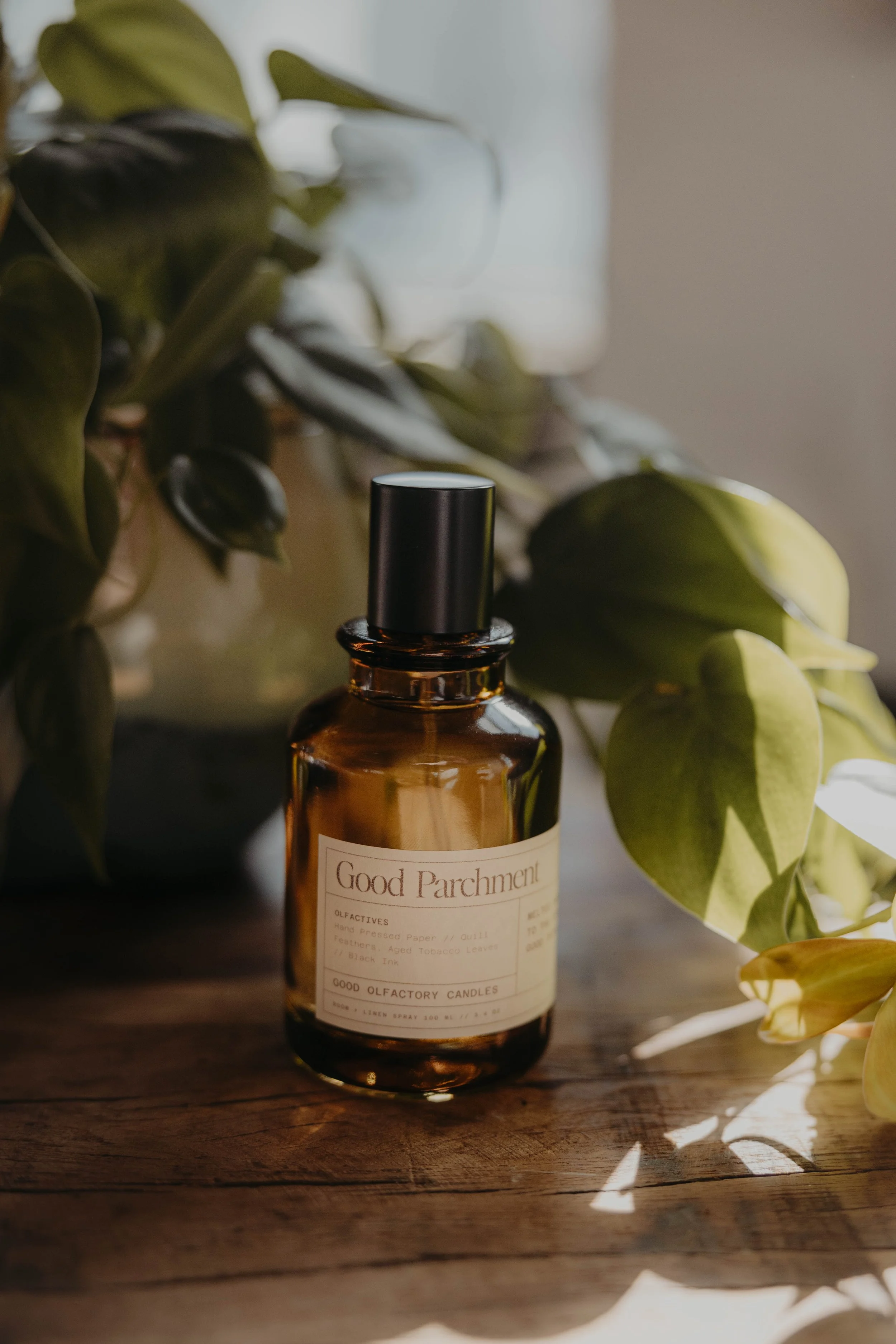 A brown glass bottle labeled 'Good Parchment' is placed on a rustic wooden surface, surrounded by green leaves with sunlight casting shadows.