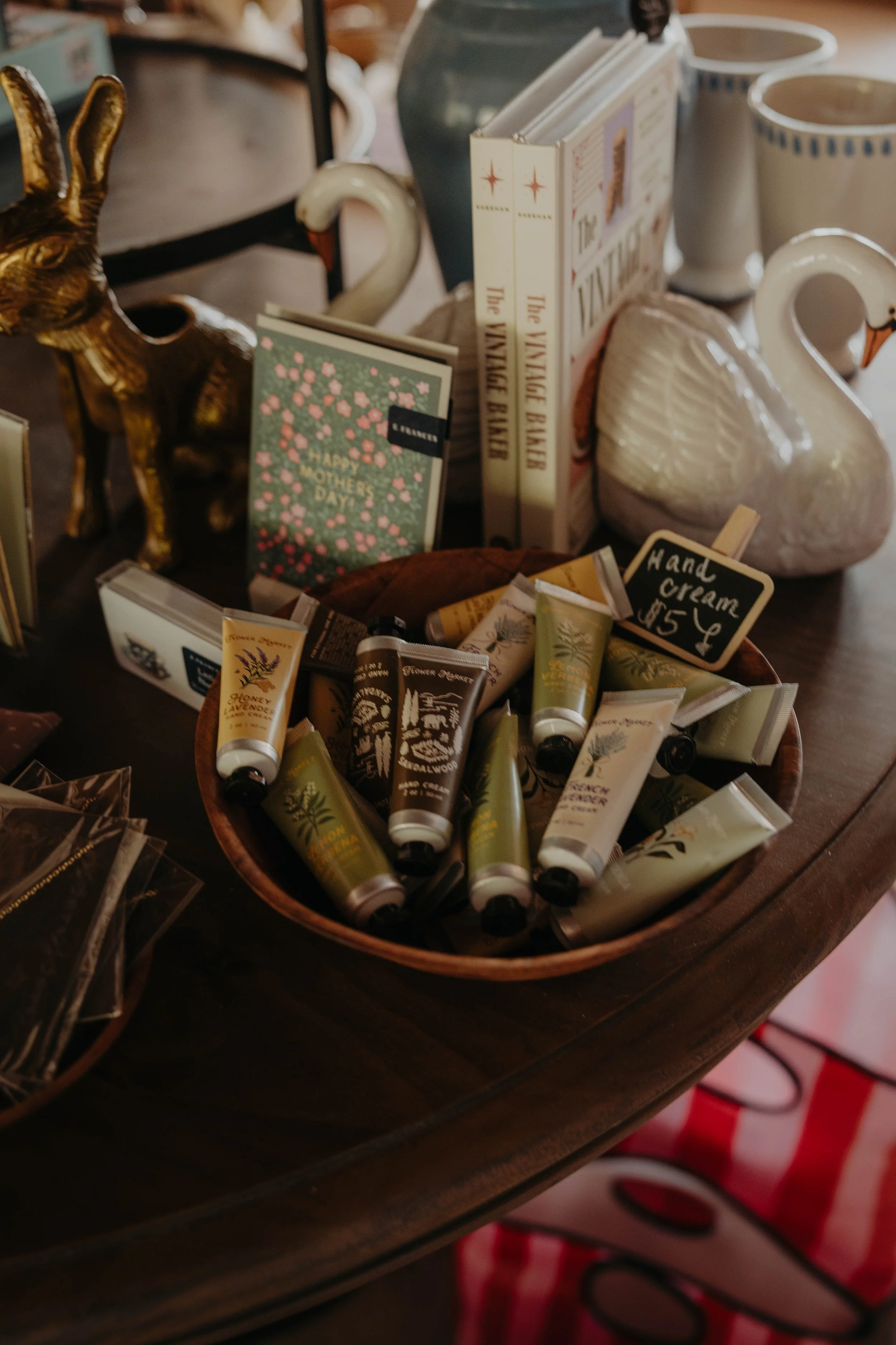 A wooden table displays various items including a golden rabbit figurine, greeting cards, books, ceramic mugs, swan-shaped containers, tubes of hand cream, and a small chalkboard sign with the price of hand cream.