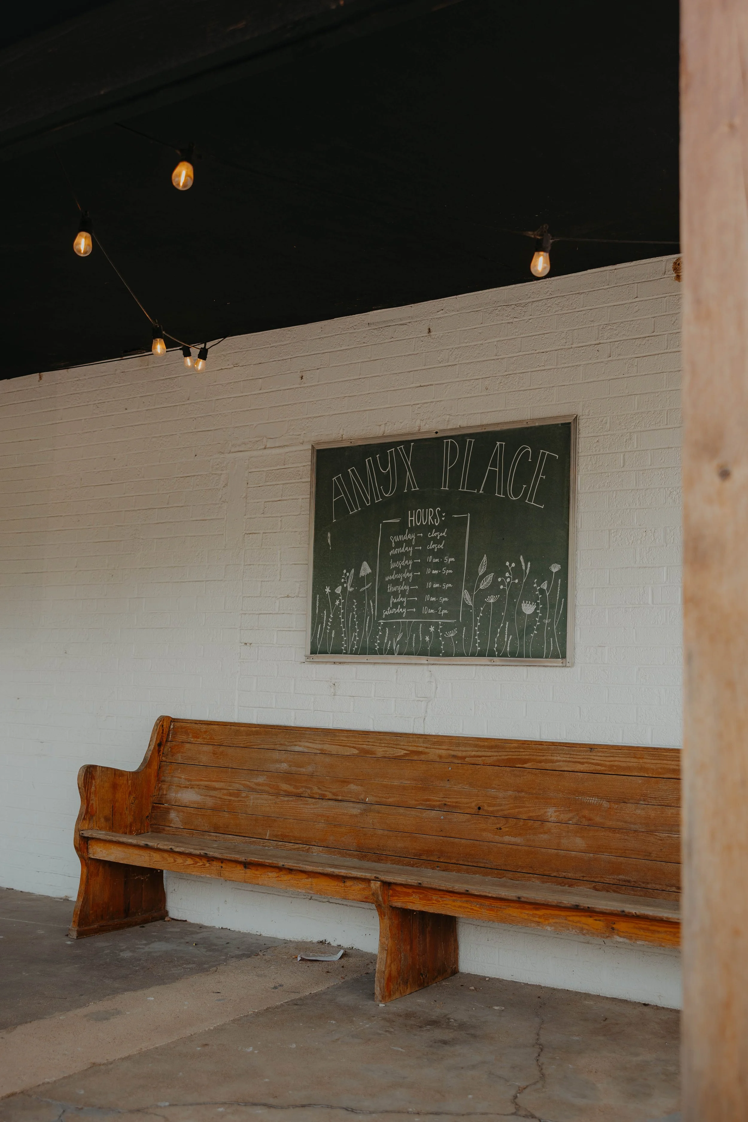 Wooden bench in front of a white brick wall with a chalkboard displaying store hours and floral illustrations, string lights hanging from the ceiling.