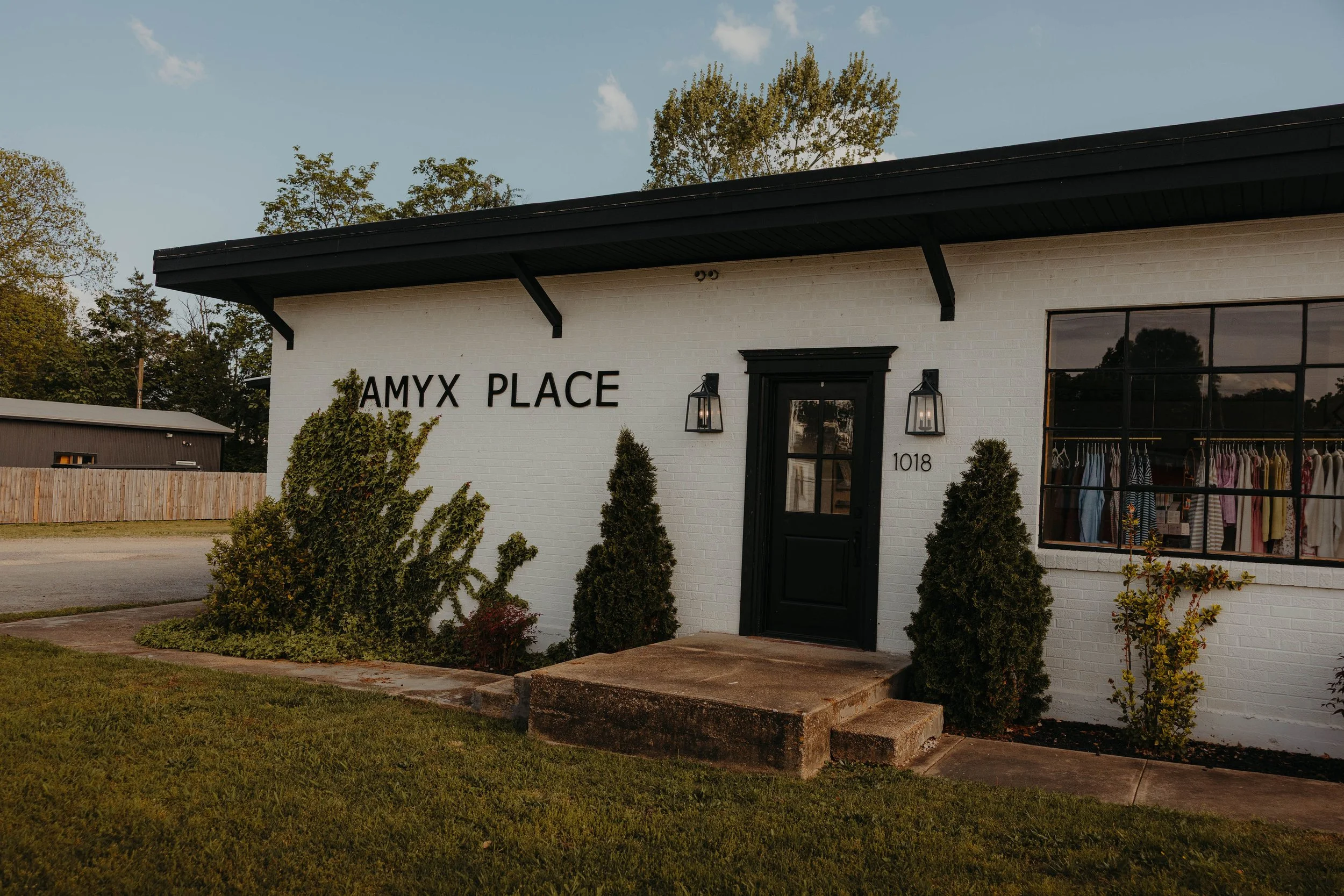 Exterior of a white brick building with black trim labeled 'AMYX PLACE,' with large windows displaying clothing, two outdoor lanterns, shrubs, and a grassy area in front.