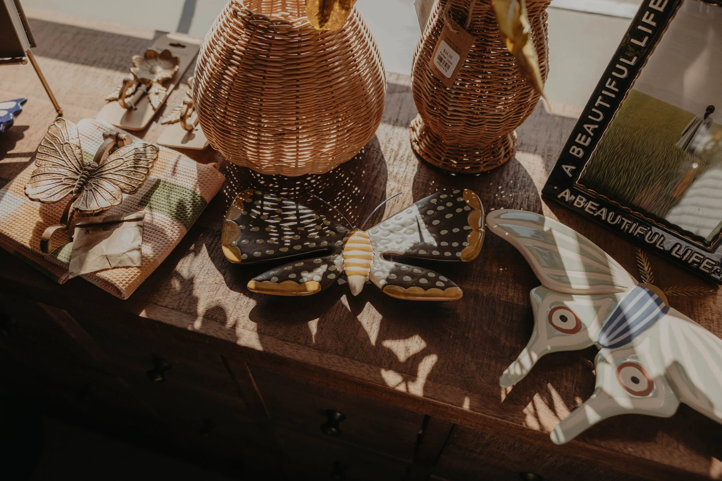 Display of various decorative home decor items, including woven vases, ceramic butterfly ornaments, a butterfly-shaped tray, and a small framed mirror with the text "A Beautiful Life" on the frame, arranged on a wooden surface.